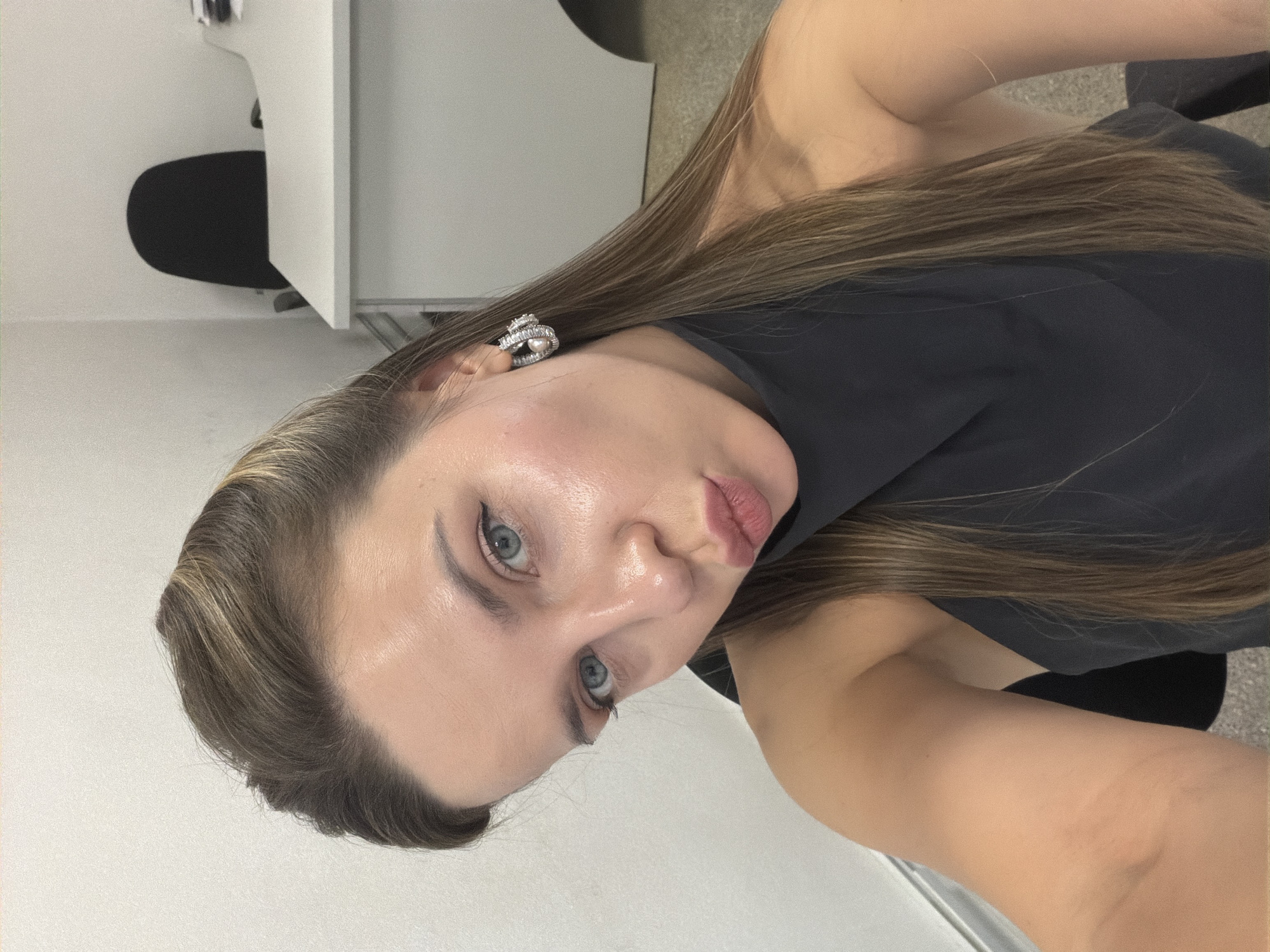 A young woman with light brown hair, blue eyes, and light skin is taking a selfie in an office setting. She is wearing a dark, sleeveless top and large, sparkly earrings. Her lips are pursed in a pout. The background shows a white wall, a black office chair, and a white desk. The lighting is bright and appears to be artificial, casting a slight sheen on her skin. The overall impression is of a casual self-portrait taken in a typical office environment. The image is from Homyel, Belarus.