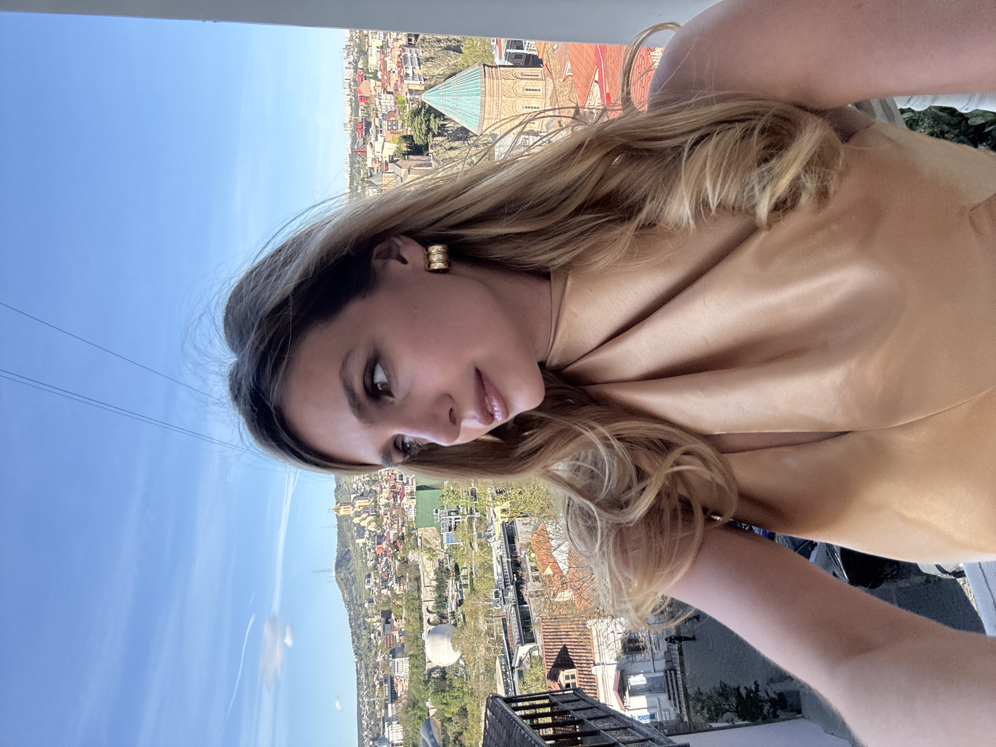 A woman with blonde hair and gold earrings is holding a cell phone to take a selfie in Tbilisi, Georgia. The background shows a cityscape with various buildings and structures. Specifically, the picture captures a view of the Sioni Cathedral, notable for its turquoise-colored dome. The sky is blue with sparse clouds. The city sprawls across the terrain, extending toward a mountain in the distance. The photo is angled, providing a partial view of the woman and her surroundings.
