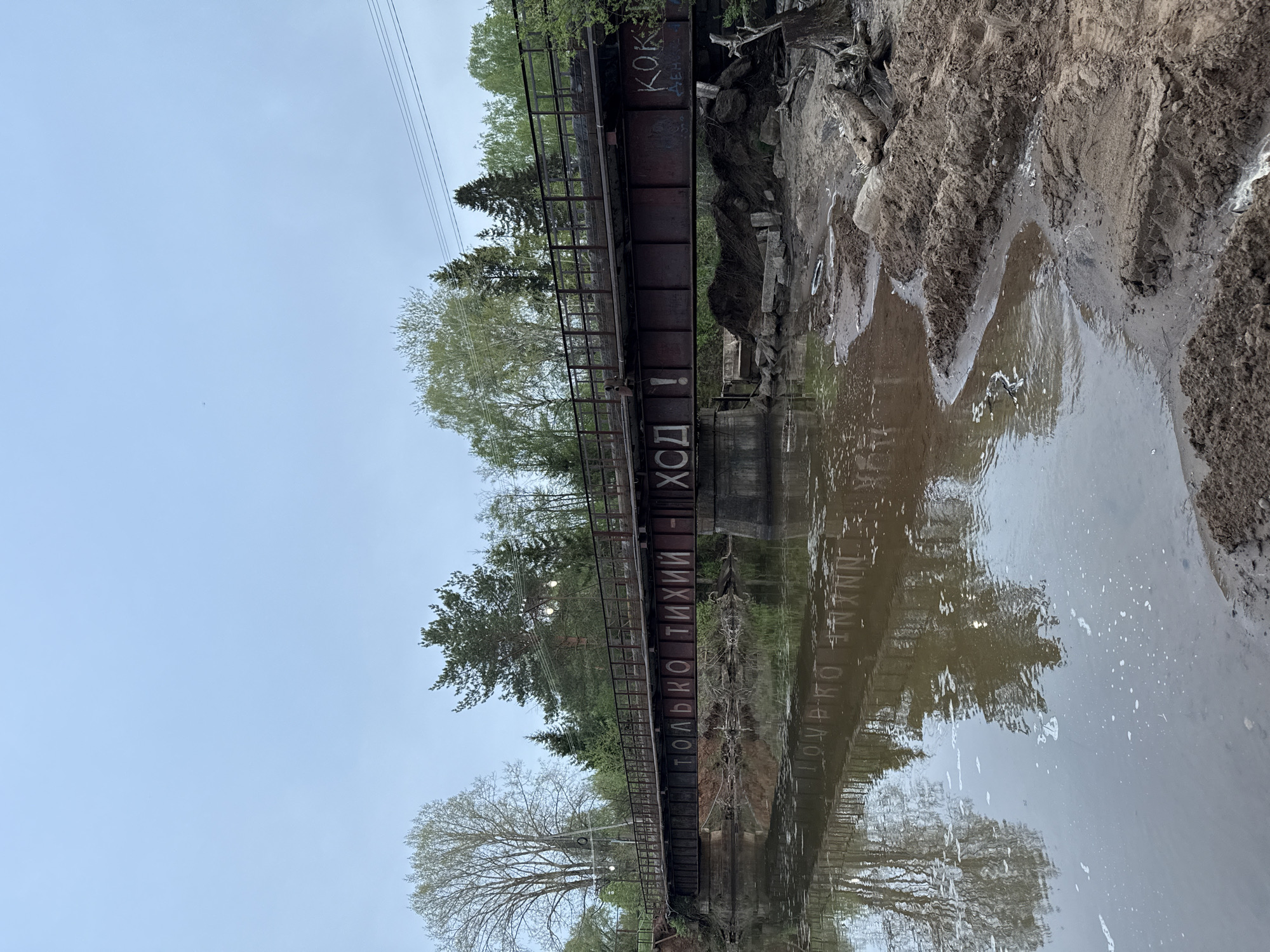 The image depicts an aged, rusted pedestrian bridge spanning a body of water. The bridge has faded Russian text painted on its side, reading "ТОЛЬКО ТИХИЙ - ХОД", and the word "КОК" is visible on the structure as well. The water reflects the bridge and surrounding foliage. The environment consists of trees, and a muddy riverbank on the right. The sky is overcast. The location is unspecified in terms of city and country.