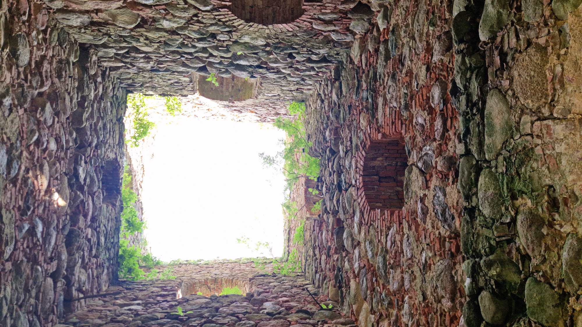 The image shows the interior of a ruined stone structure, likely a castle or fortification, looking upwards towards an open sky. The walls are constructed from rough-hewn stones and bricks, some of which are covered in moss and vegetation. There are arched openings visible in the walls, and the floor appears to be made of rubble. The scene is characterized by a sense of age and decay. This is a view of the ruins of the Embūte castle mound and manor house, located in Embūte, Latvia.