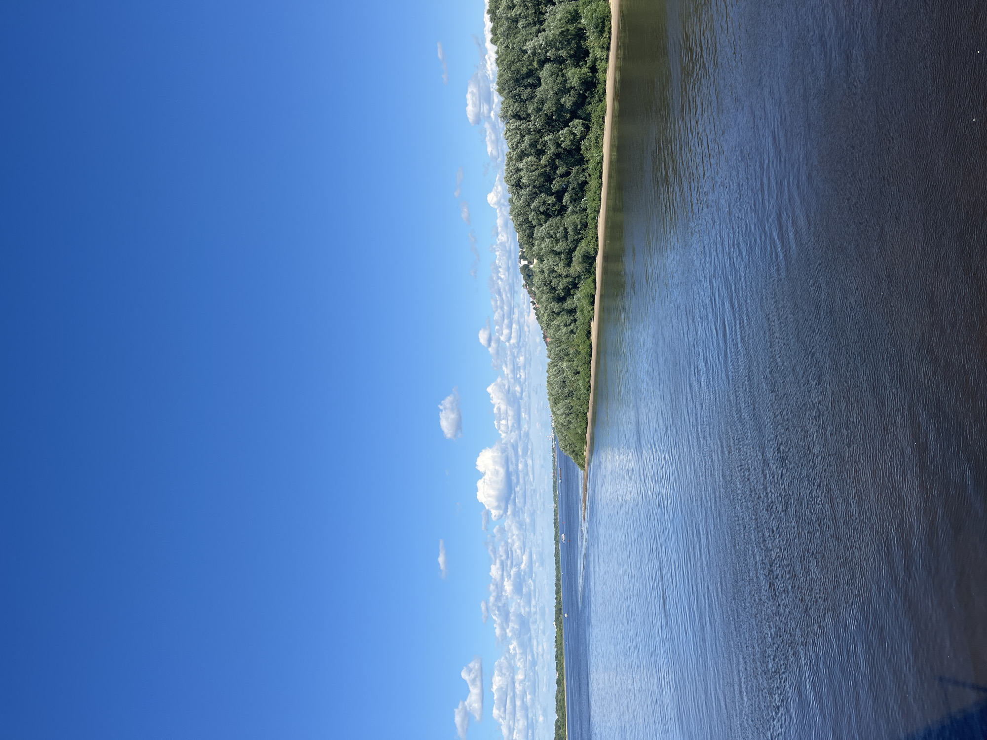 This image shows a wide, dark blue river in Nizhny Novgorod, Russia, under a bright blue sky dotted with white, fluffy clouds. The river is calm with gentle ripples. On the right bank, a dense, green forest covers a hillside. The trees are lush and full, and the shoreline is a light brown sand. In the distance, across the river, the far bank is also lined with trees, and hints of buildings can be seen. Several small boats are visible on the water in the distance. The overall scene is one of natural beauty and tranquility on a clear, sunny day.