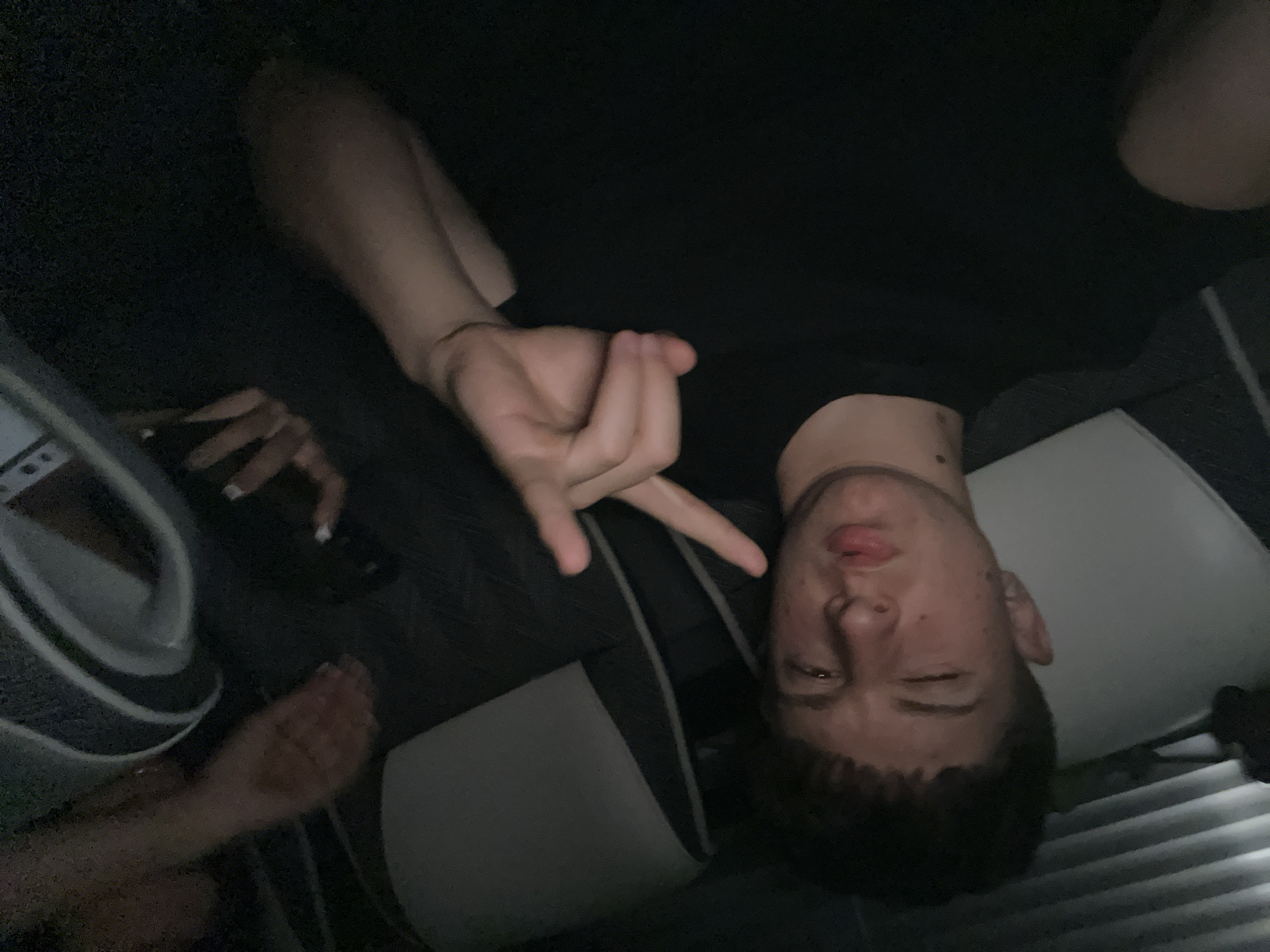 A male individual is seen indoors, angled slightly, making a gesture with his hand. He has short, dark hair and is wearing a dark shirt. Another person's hand is visible holding a dark object, possibly a phone. Beside them is an object which appears to have an outer ring. The environment seems dimly lit. Blinds are visible. The location is not discernible from the image.