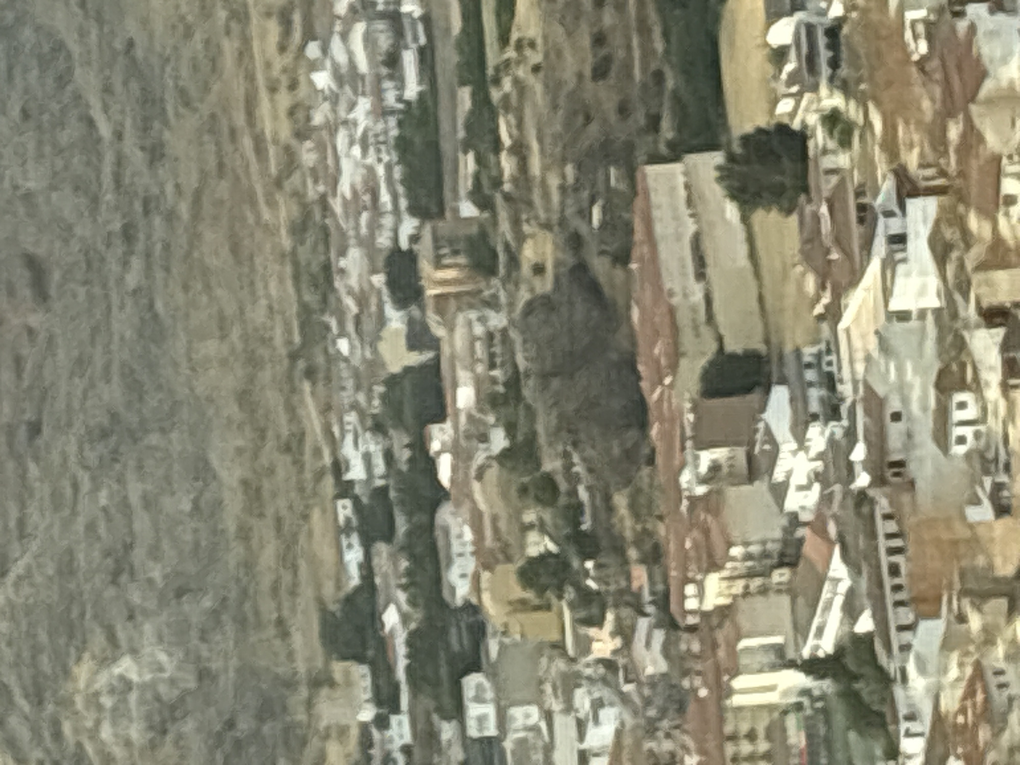 The image shows a panoramic view of the city of Loja, Spain, characterized by its hilly terrain and dense urban fabric. The architecture features a mix of white-washed buildings with terracotta roofs, typical of Andalusian villages. The city is nestled at the foot of a rugged, mountainous landscape, with the textured slopes of the hills dominating the upper portion of the frame.
The foreground and midground reveal a cluster of houses, some with multiple stories, arranged along winding streets and alleys that ascend the hillsides. The buildings are tightly packed, creating a sense of a dense, ancient settlement. There are indications of greenery scattered throughout the urban area, including trees and small patches of vegetation.
In the midground, a larger, light-colored building with a prominent, possibly arched, structure stands out, suggesting a significant public building or landmark. The overall impression is one of a historic town integrated into a dramatic natural setting. The quality of the image is somewhat blurred, possibly due to the distance from which it was taken or the photographic conditions, which imparts a painterly or impressionistic quality to the scene. There are no discernible people or specific actions visible in the image.