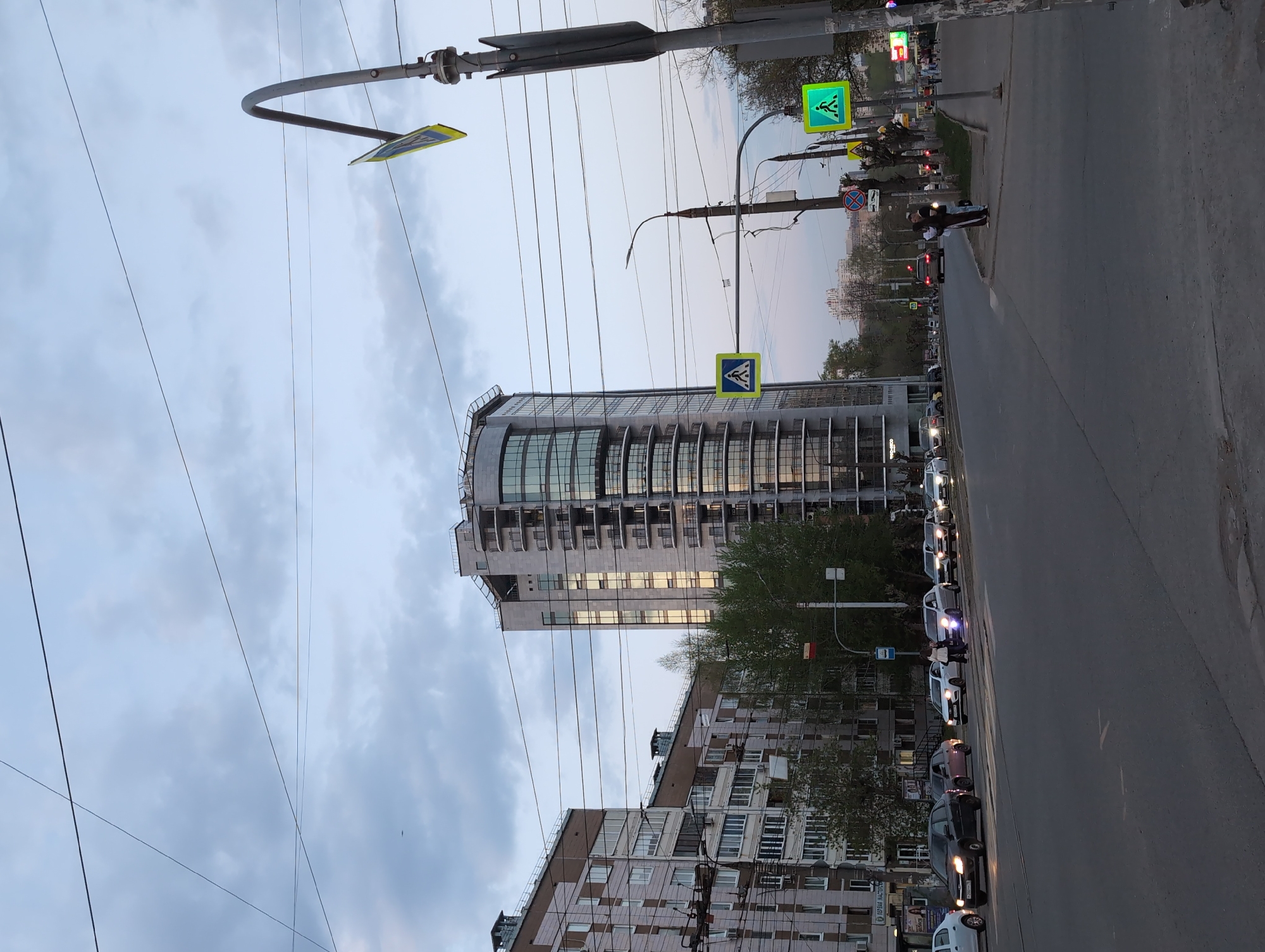 The image shows a street scene in Izhevsk, Russia. The foreground consists of a wide, dark asphalt road with a line of parked cars on the right side. A pedestrian is crossing the road between the vehicles. In the midground, there are several apartment buildings and a taller, modern office building with a curved facade. Power lines crisscross the sky above the buildings. Street signs and traffic signals are visible, including a pedestrian crossing sign with a green pedestrian symbol. The sky is overcast with a mix of light gray and blue clouds.