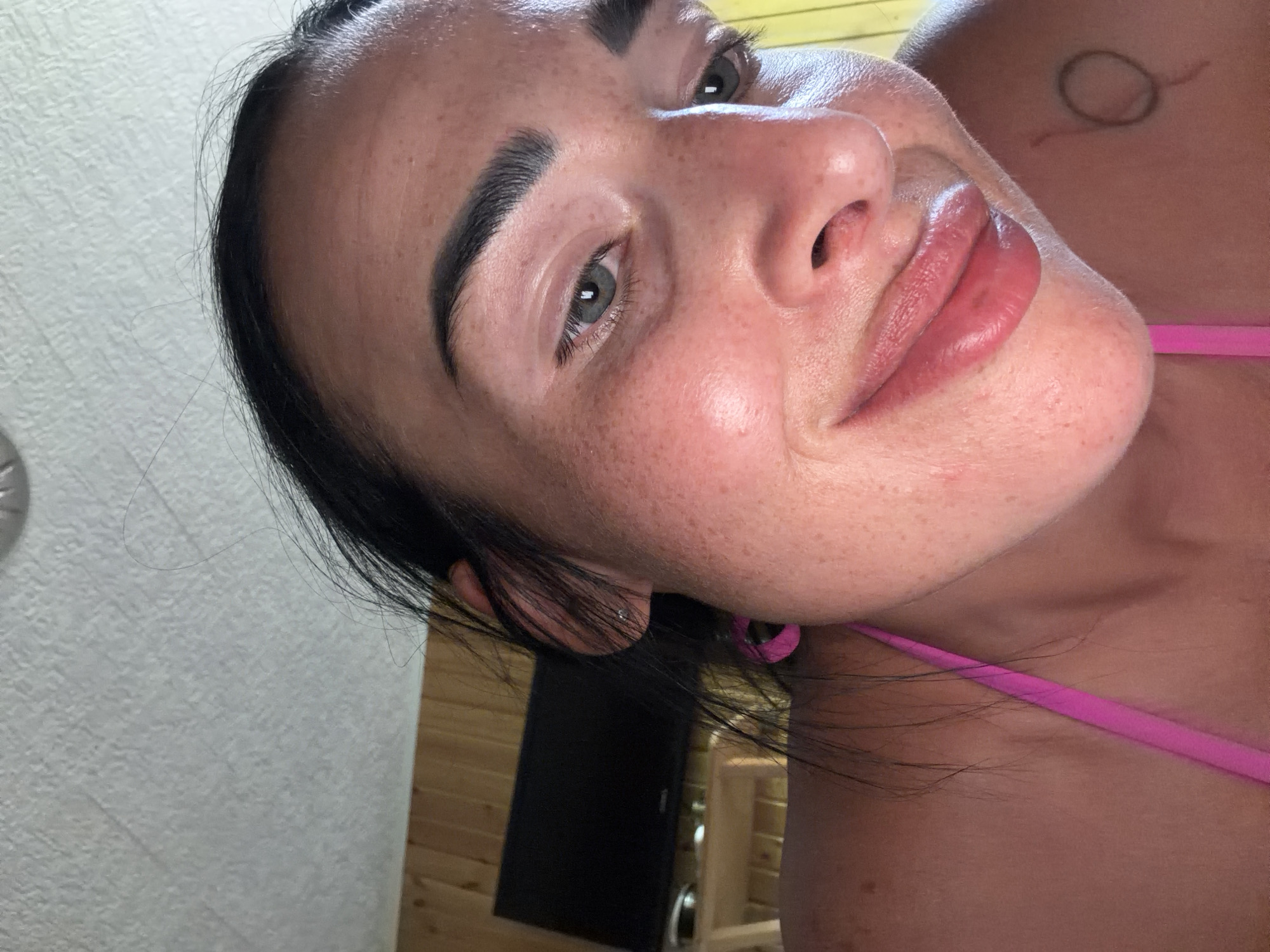 The image shows a close-up, angled selfie of a woman. She has dark hair and light skin with freckles. Her eyes are open and she is looking towards the camera. She is wearing a pink bikini top. In the background, there is a textured wall and wooden paneling, suggesting an indoor setting, possibly a room with a sauna or spa-like aesthetic. There is a faint tattoo on her upper back, appearing as a circular design. The lighting suggests it is daytime. The woman's expression is neutral. There is no visible text in the image.