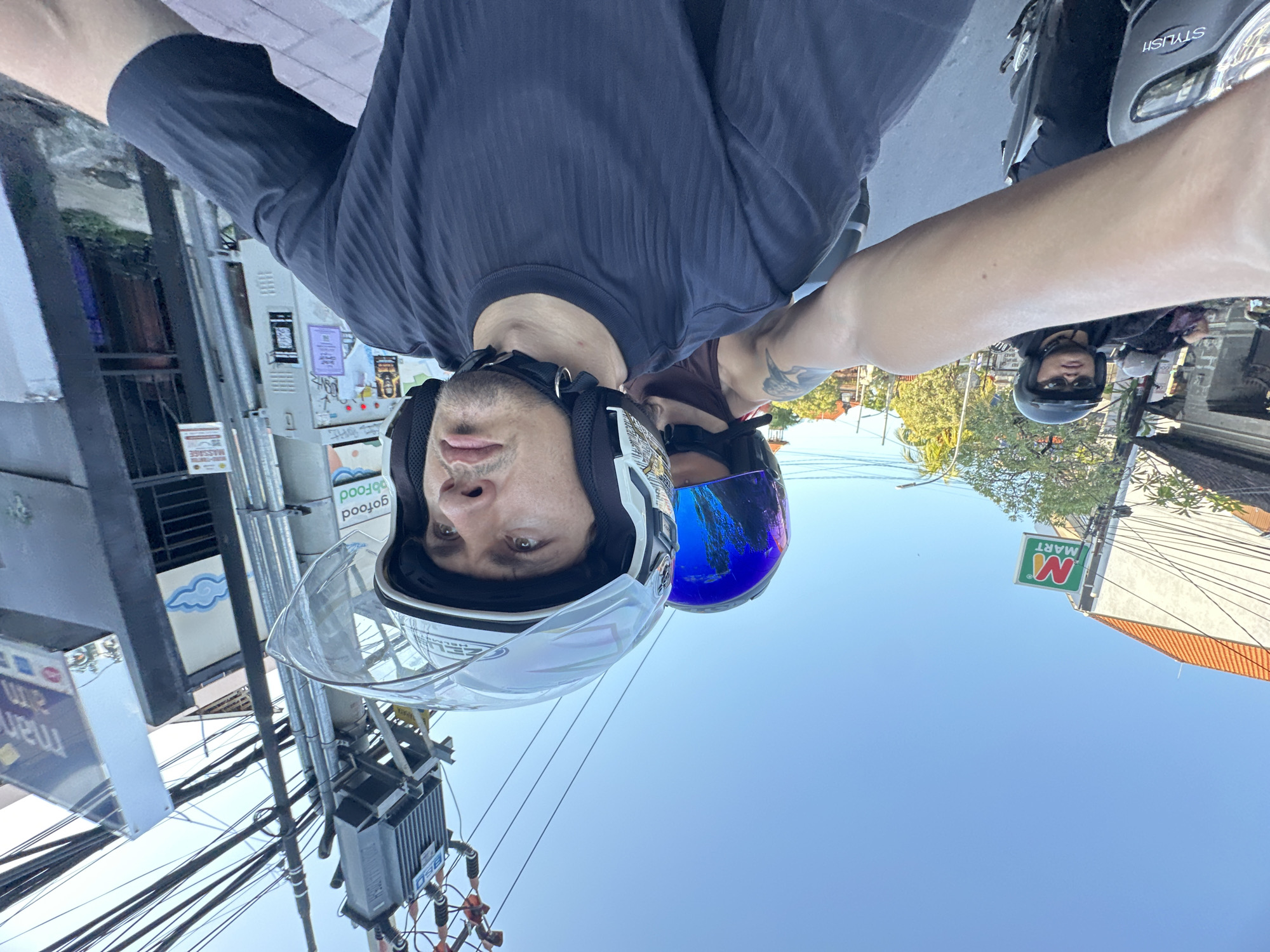 The image shows a selfie taken by a person wearing a white helmet while riding a scooter. Another person with a blue helmet is beside them. In the background, there's an "M Mart" sign, street power lines, and other scooters with riders. The location appears to be North Kuta, Indonesia.