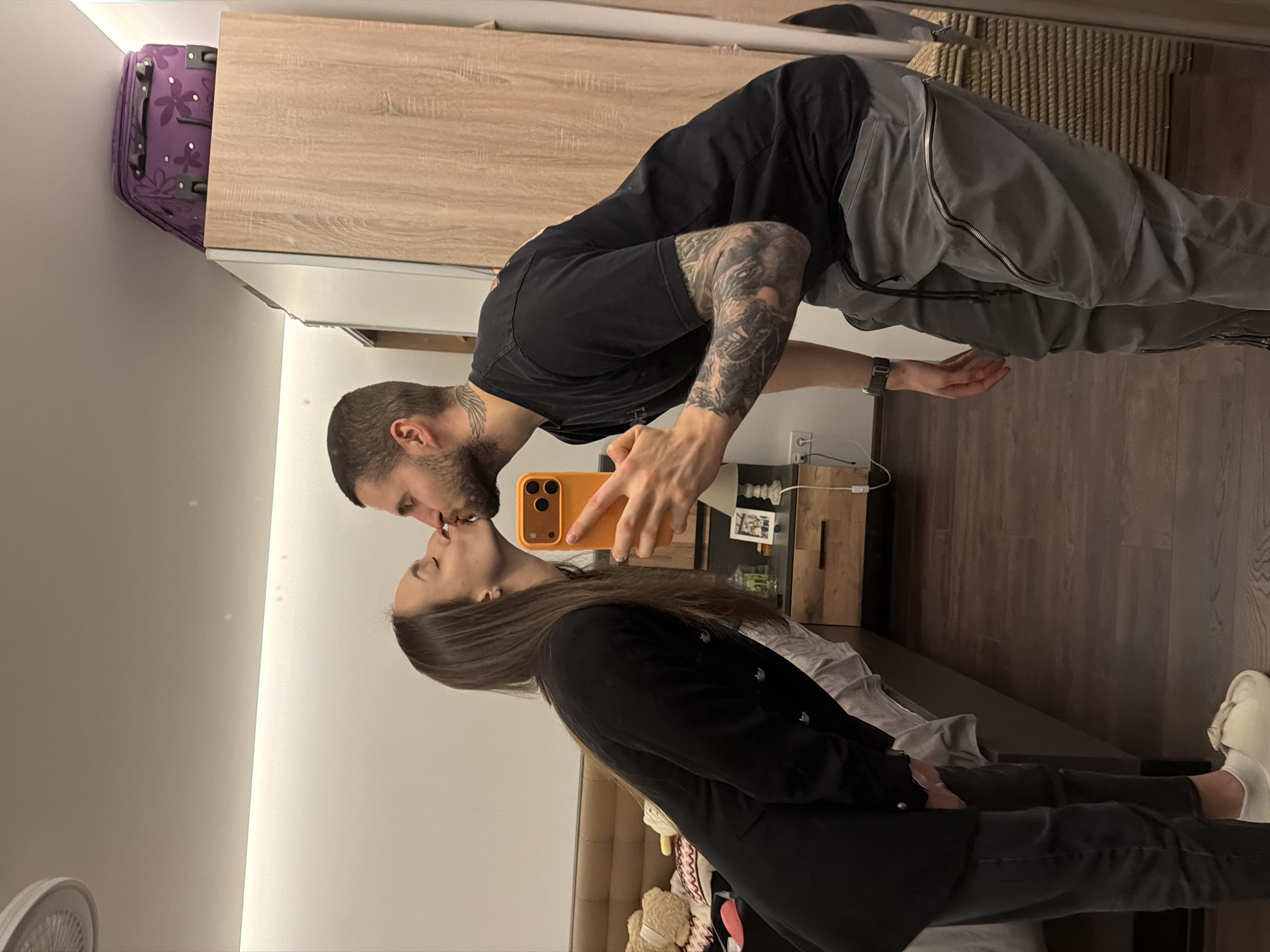 A photograph depicts a man and a woman kissing while the man holds a phone in front of them. The man has a beard and tattoos on his arm. He is wearing a black t-shirt and grey pants. The woman is wearing a black jacket. The background includes a bed, a wooden dresser, and a purple suitcase. The location cannot be determined to be in the city of Sofia, Bulgaria, based on the visual information.