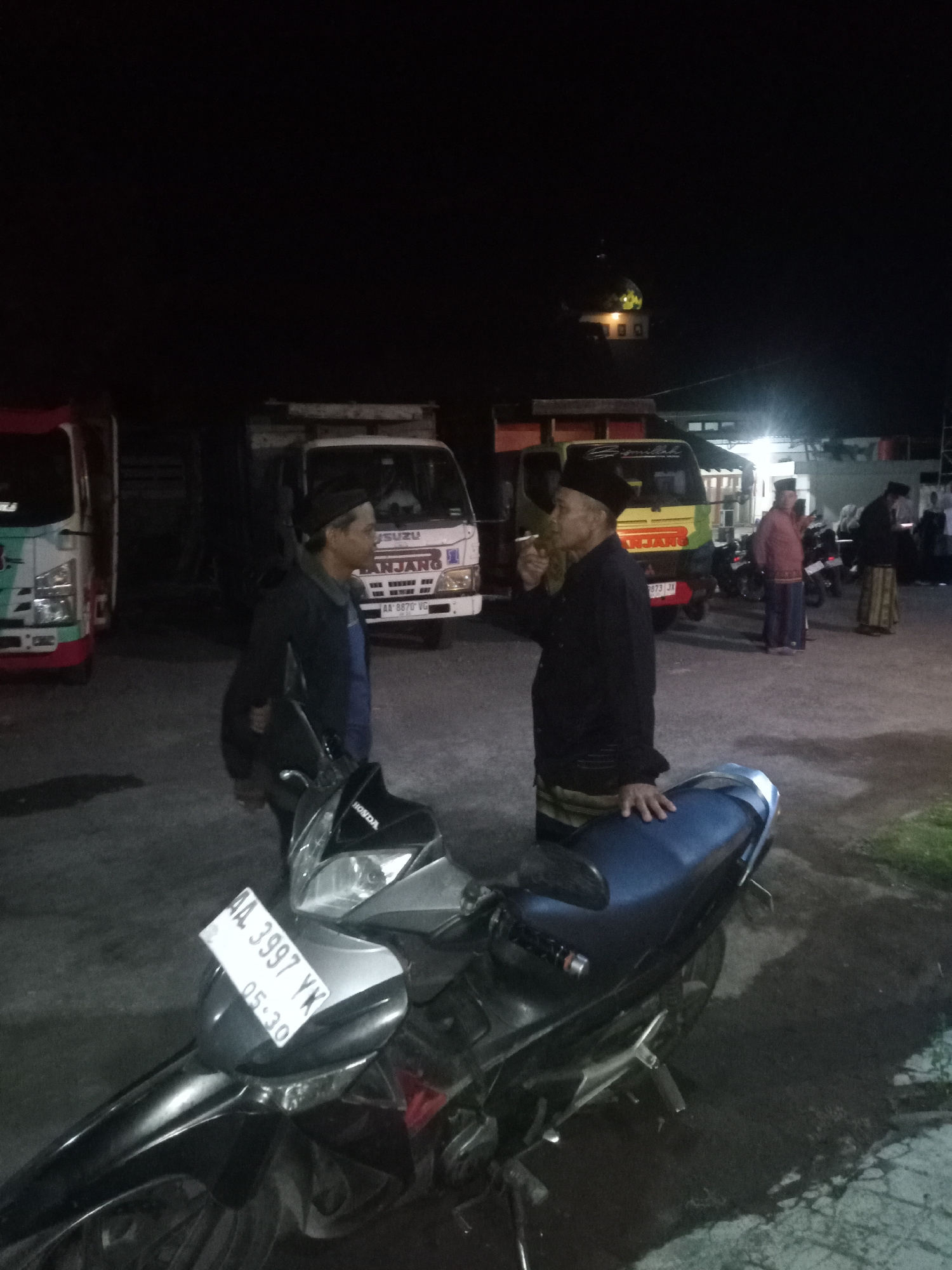The image depicts a nighttime scene in what appears to be an outdoor area, possibly a parking lot, in Majaksingi, Indonesia. In the foreground, a black motorcycle is parked, with its license plate clearly visible, reading "AA 3997 YK 05-30". Two men are the primary subjects in the mid-ground. The man on the left is wearing a dark blue jacket and a black cap, with his hands clasped in front of him. He is facing right, looking towards the other man. The man on the right is wearing a dark long-sleeved shirt, a black cap, and a sarong. He is holding a cigarette in his right hand and appears to be smoking. He is facing left, engaged in conversation or interaction with the other man.
In the background, several vehicles are parked, including two trucks. One truck is white with "ISUZU" and "LANJANG" written on its side, and its license plate is "AA 8870 VG". Another truck, partially visible on the left, is white with red accents and has the "CMC" logo on it. Further in the background, other people are visible, some of whom are wearing traditional Islamic attire, suggesting a possible cultural or religious gathering. A building with a domed roof, possibly a mosque, is visible in the far background, illuminated by lights. The overall environment is dark, indicating it is nighttime, with artificial lighting from the vehicles and structures casting shadows.