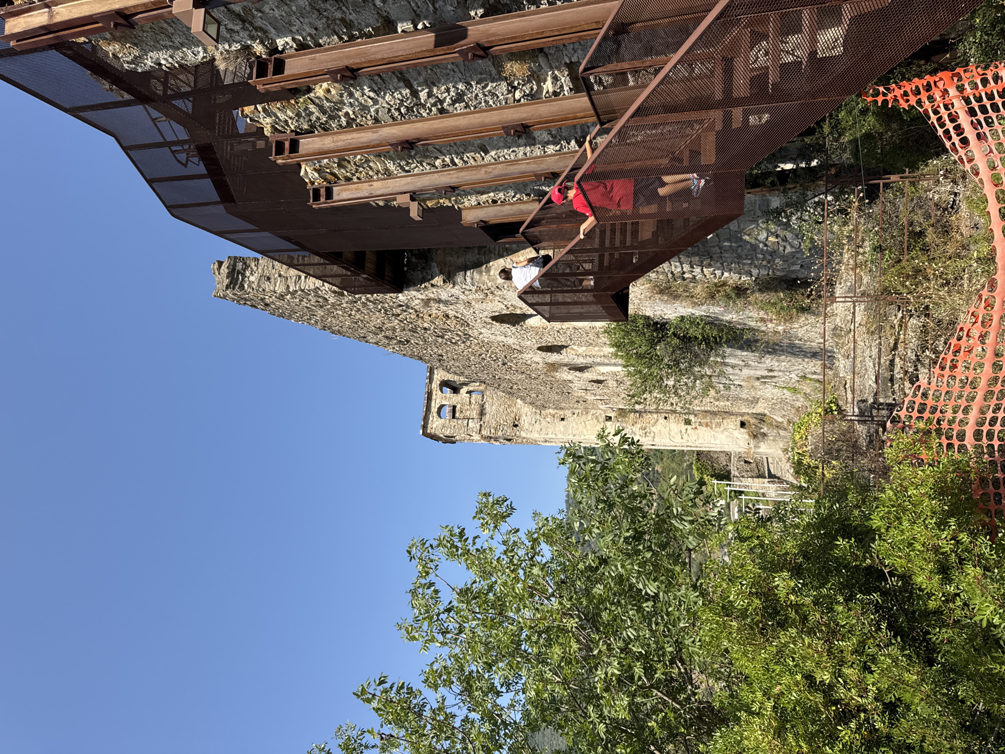 An exterior, low-angle shot shows a weathered stone structure resembling a castle or ancient ruin, with a modern, rust-colored metal staircase and walkway attached. The sky is a clear, bright blue. To the left, a vibrant green tree with dense foliage partially obscures the stone building. The stone structure has a tower-like section with arched openings. The metal staircase, made of grated material, winds its way up the side of the stone building, supported by vertical steel beams. Two people are visible on the staircase: a person in a white shirt and dark pants is higher up and further away, while a child wearing a red cap and shorts is closer to the foreground, ascending the stairs. The environment appears to be a historical site with some areas cordoned off by orange construction netting in the foreground. The scene suggests a warm, sunny day. The location is the Doria Castle in Dolceacqua, Italy.