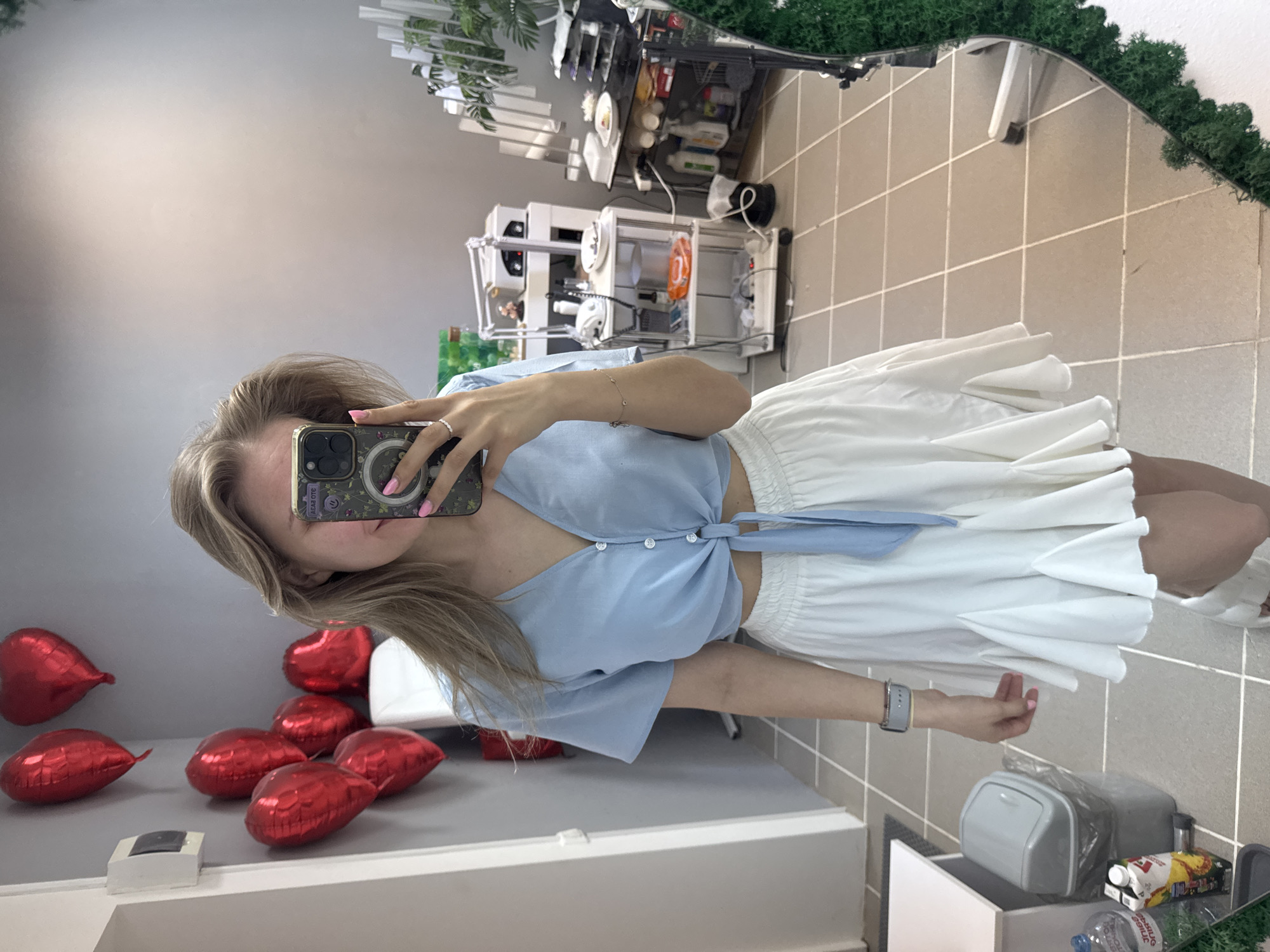 A young woman is taking a selfie in a mirror. She has blonde hair and is wearing a light blue tied top and a white skirt. In the background, there are red heart-shaped balloons and what appears to be a 3D printer on a shelf. The room is tiled, and a decorative green element is present around the mirror. The location of Izhevsk, Russia cannot be confirmed from this image.