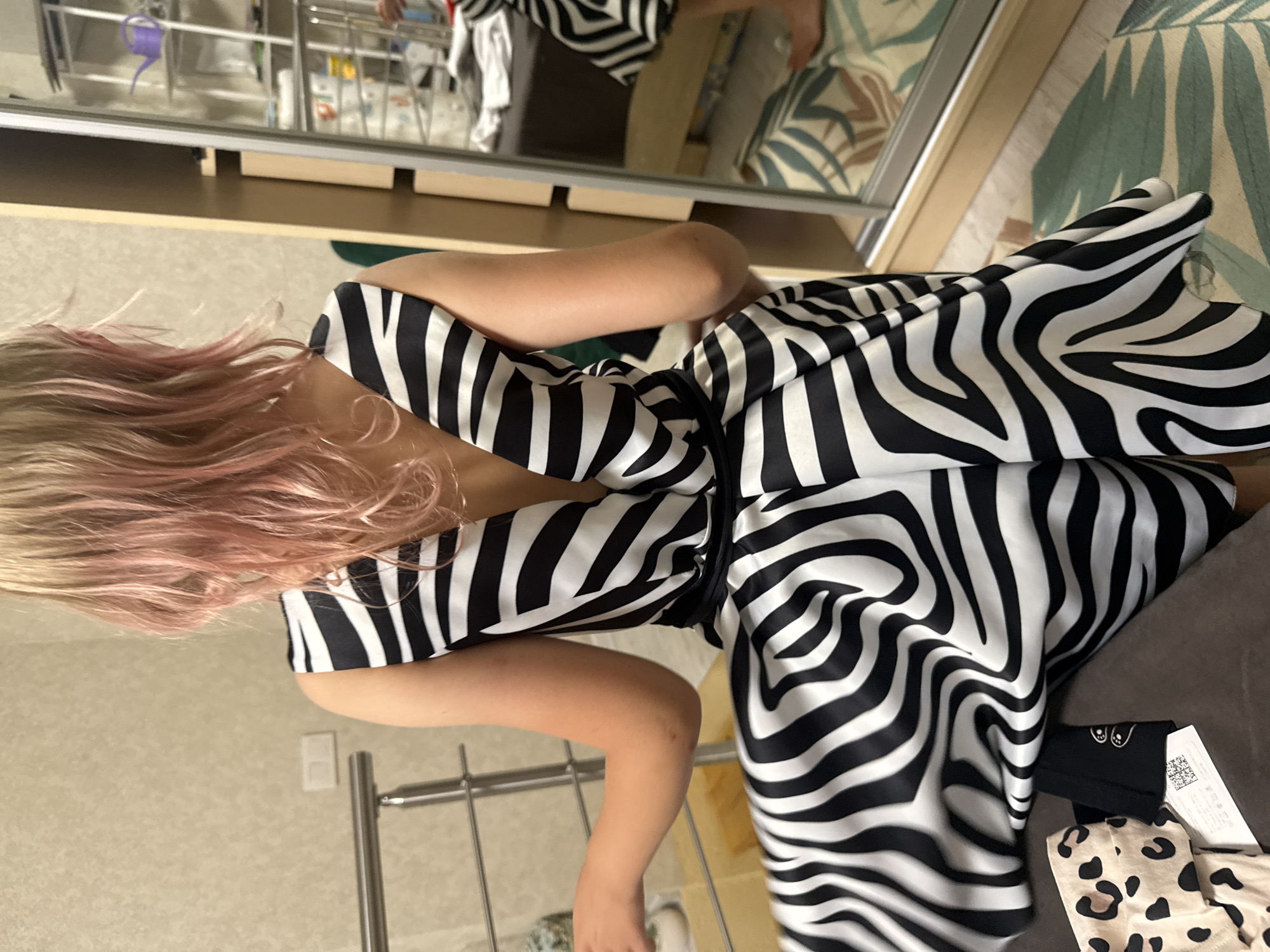 In the city of Unknown, Unknown, a person is photographed from behind, wearing a black and white zebra-print dress. The dress has a deep V-neckline in the back and a flowing skirt. The person has blonde hair with pink tips. They are standing next to a tall mirror wardrobe, with a small shelf unit visible inside. A metal railing is to the left of the frame, and a patterned rug is on the floor. There are also a few items of clothing on a brown surface in the foreground, including a leopard-print item. The overall scene suggests an indoor setting, possibly a bedroom or dressing area.