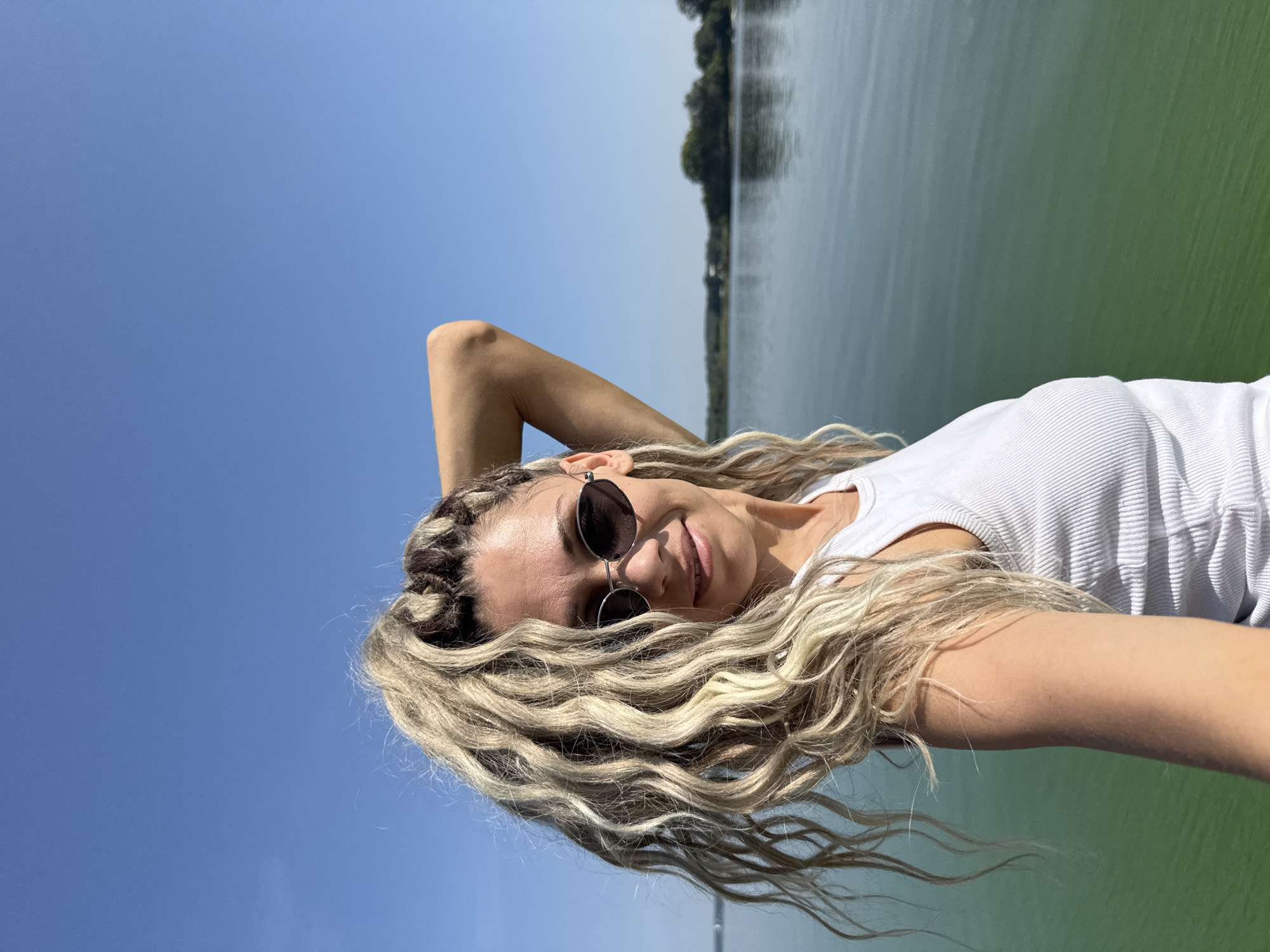 The image is a portrait of a woman with blonde dreadlocks standing outdoors. She is wearing sunglasses and a white tank top. Her right arm is raised behind her head. The background consists of a body of water with a tree-lined shore in the distance, under a clear blue sky. The location is stated to be in the city of Rusanovo, Russia.