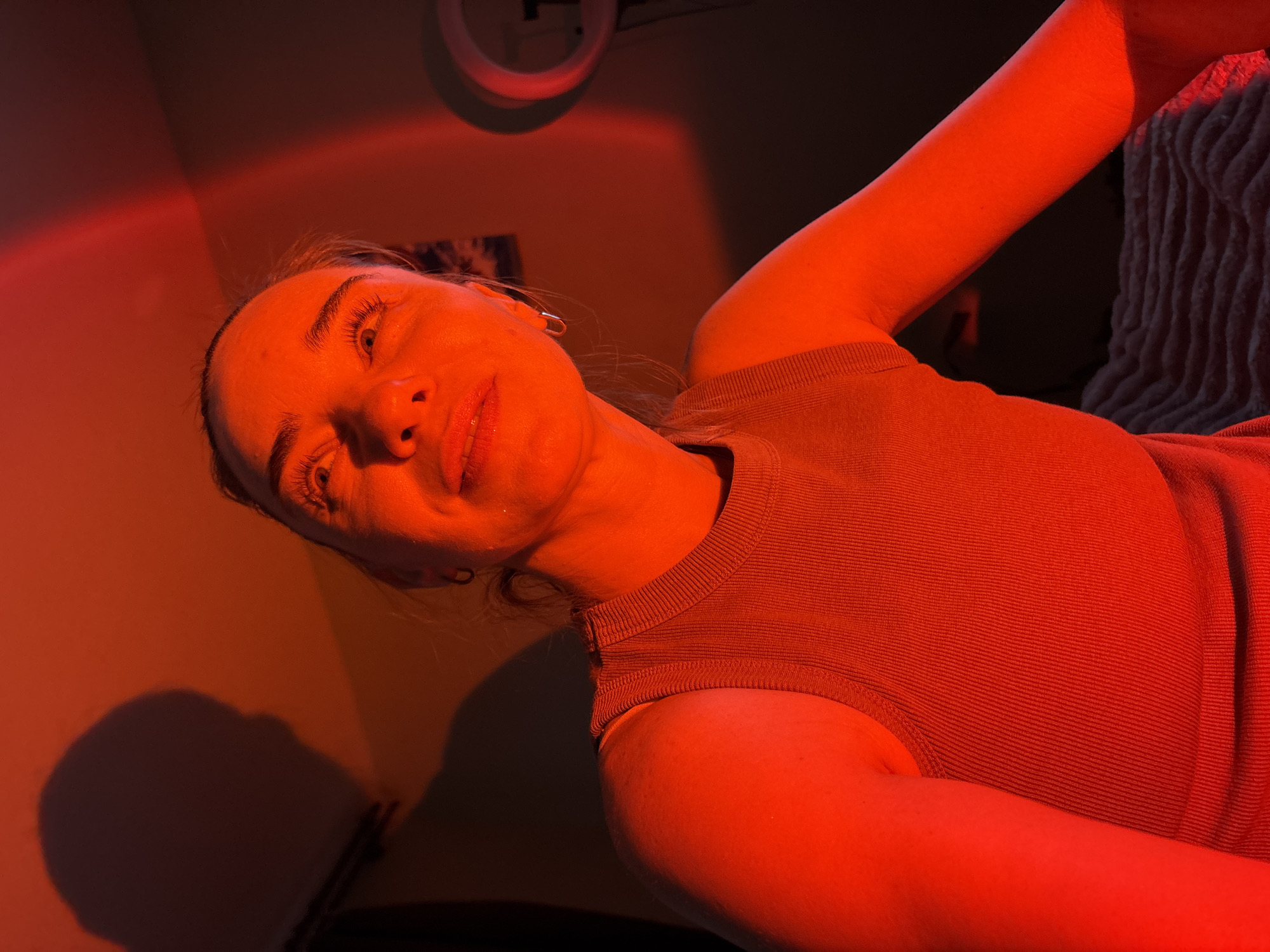 The image is a close-up, low-angle, portrait-style photograph of a young Caucasian female illuminated by red light. She is positioned in the center of the frame, looking upwards and to her right with a slight smile. Her face shows signs of makeup, including mascara and lip gloss. She is wearing a ribbed, sleeveless tank top in a muted, reddish-brown hue. A faint shadow of her head and shoulders is visible on the wall behind her, cast by an unseen light source. To the right of the frame, a round, white object with a dark center is partially visible, possibly a ring light or a decorative item. The background consists of a wall with a subtle gradient from a dark reddish-brown to a lighter, warmer tone. A hint of a textured surface, possibly a blanket or bedspread, is visible in the lower right corner. The overall lighting creates a dramatic and moody atmosphere. No specific location in a city of "Unknown, Russia" can be determined from the visual information provided.