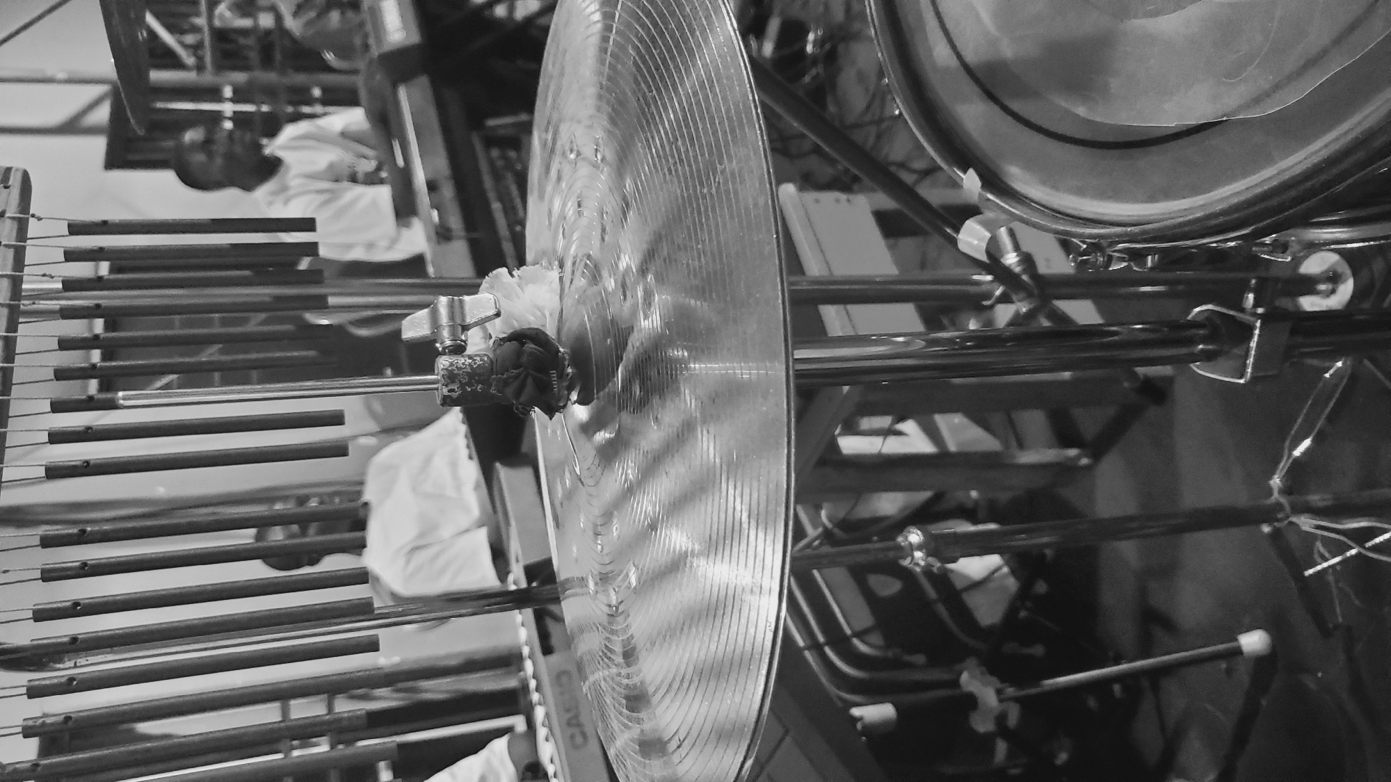 The image captures a close-up view of a cymbal on a drum stand, with several other musical instruments and individuals in the background. The cymbal is metallic and appears to be in use, with visible concentric rings. Above the cymbal, a set of vertical metal bars, likely part of a percussion instrument like a glockenspiel or chime bar, hangs from a wooden frame. In the middle ground, two men are visible, both wearing white shirts, seated in front of keyboards, suggesting a live music performance or rehearsal. One keyboard prominently displays the "CASIO" logo. The overall environment appears to be an indoor space, possibly a church hall or a performance venue, with a concrete floor and visible electrical wiring and equipment. The lighting is somewhat dim, contributing to the black and white aesthetic. The image does not provide specific clues about the precise location within Jalingo, Nigeria, beyond it being a setting for a musical event.