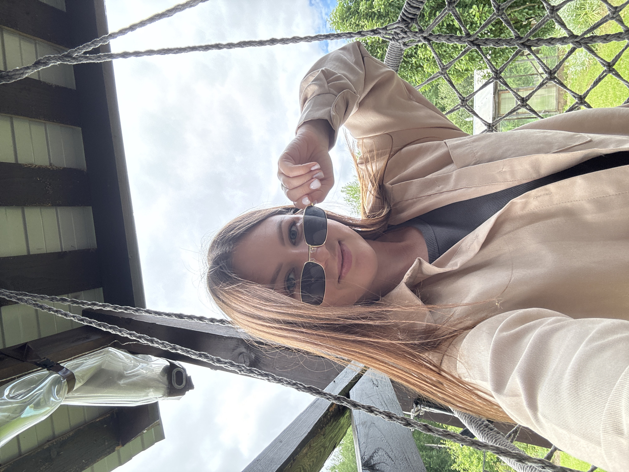 This is a selfie of a young woman taken outdoors. She is wearing a beige trench coat over a dark grey top and black sunglasses. Her long, brown hair is flowing. She is holding her sunglasses up with her right hand. The setting appears to be a wooden structure, possibly a treehouse or an elevated platform, with thick ropes forming a net-like structure overhead. In the background, green trees and grass are visible, suggesting a natural environment. The sky is cloudy. A clear plastic bottle, possibly a water bottle, is attached to the wooden structure with dark tape on the left side of the frame. The image is taken from a low angle, looking up at the woman. The orientation of the image is sideways. The location is specified as Ponazyrevo, Russia. There is no visible text in the image. The overall mood seems casual and relaxed.