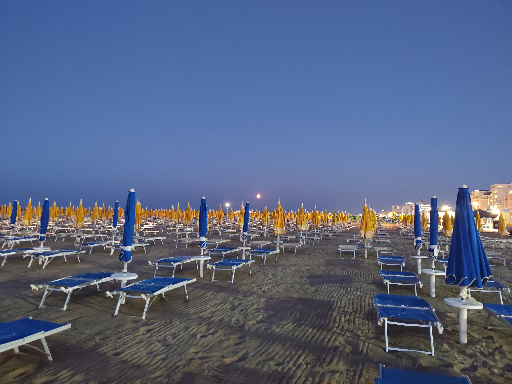 The image depicts a deserted beach in Rimini, Italy, at twilight. Rows of empty blue and yellow beach umbrellas and lounge chairs stretch across the sand, which is rippled with patterns from the receding tide or wind. In the background, lights from buildings along the coastline are beginning to illuminate. The sky is a deep, clear blue, with a full moon visible near the horizon. There are no people present in the scene.
