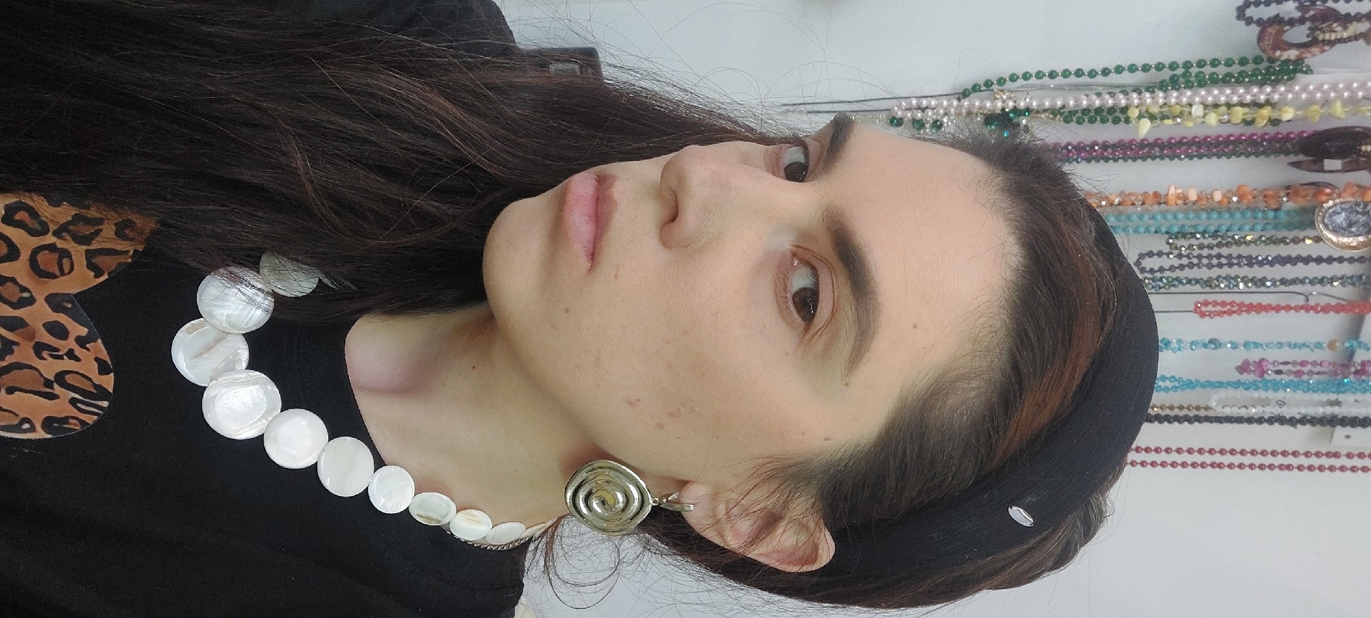 A person is shown in an interior setting. The individual has long, dark hair, a fair complexion, and is wearing a black headband. A necklace of white, circular beads adorns their neck, and a spiral-shaped earring is visible on one ear. The person is dressed in a black shirt with a leopard print patch on the shoulder.
In the background, several beaded necklaces are displayed on a wall, suggesting the location may be a jewelry store. The store is possibly located in Novosibirsk, Russia.