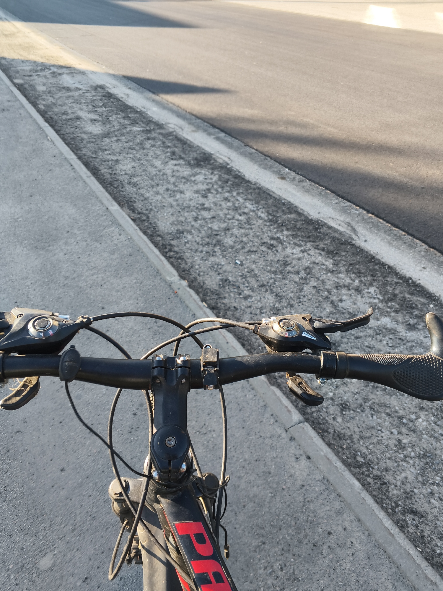 This is a first-person perspective image taken from a bicycle, looking forward along a road. The handlebars of the bicycle are prominently featured in the foreground, with black grip tape on the right side and gear shifters visible on both sides. The bicycle frame, black with red lettering that partially spells out "PA", is visible below the handlebars. The road appears to be asphalt, with a distinct concrete curb and a gravel shoulder to the left. The sun is casting shadows across the road, suggesting it is either early morning or late afternoon. The overall environment is an urban or suburban roadside setting. There are no people or animals visible in the image. The location is identified as Mokshan, Russia.