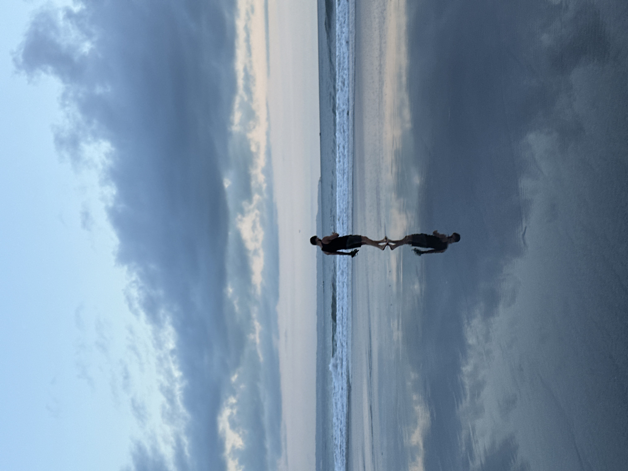 A low-angle, landscape-oriented photograph captures a beach scene in North Kuta, Indonesia, at dusk or dawn. The sky, a gradient of blue and grey clouds, takes up approximately half of the image. Below, the wet sand reflects the sky, creating a mirrored effect. A single, dark-skinned person, likely a male, is walking on the beach near the water's edge. The person's reflection is also visible in the wet sand. The ocean is represented by a strip of white waves crashing against the shore. The overall impression is one of tranquility and solitude.