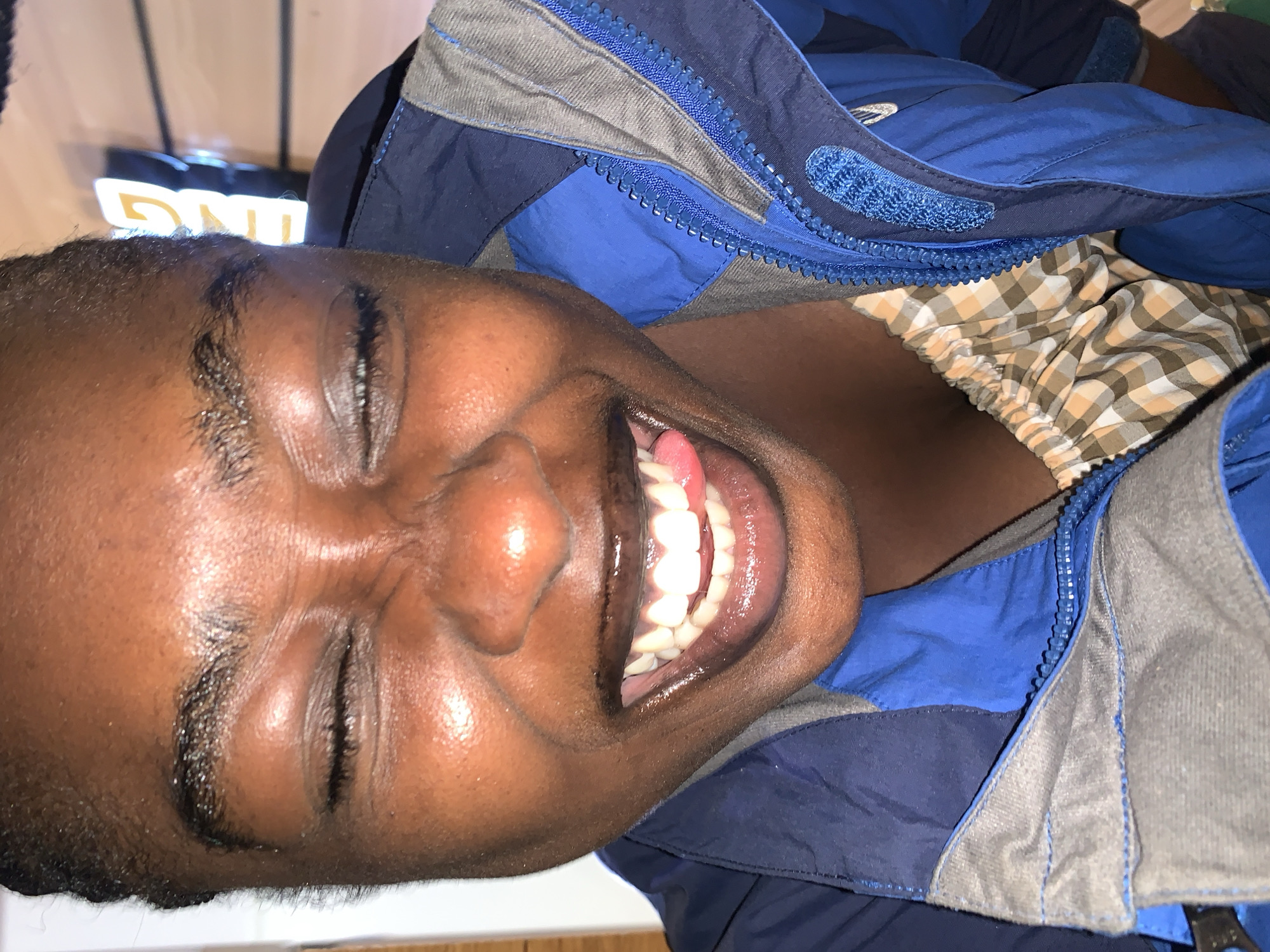 The image shows a close-up of a young Black person with their eyes squeezed shut and a wide smile, sticking their tongue out slightly. They are wearing a blue jacket with gray accents and a checkered undershirt. The background is blurred, but appears to be indoors with some lighting and possibly signage, suggesting a commercial establishment. There is no discernible information in the image to indicate the location is Gura Top, Nigeria.