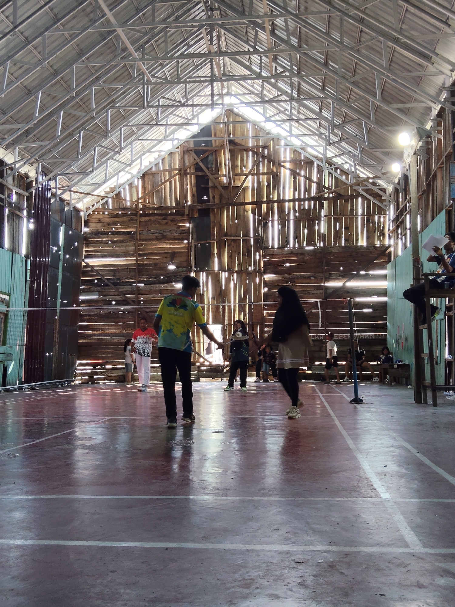 The image depicts the interior of a rustic badminton court within a wooden structure, likely in Lori, Indonesia. Sunlight streams through gaps in the corrugated metal roof and the weathered wooden walls, creating stark vertical lines of light. The court floor is a dark, reflective surface with white lines marking boundaries. Several individuals are present, engaged in various activities. In the foreground, a person in a yellow and blue shirt faces away from the camera, while another person in a dark hijab and light-colored skirt walks away. In the mid-ground, a person in a red and white shirt stands near a child in a light-colored outfit. Other figures are visible further back, some seated and observing. Badminton nets are set up on the court, and a judge's chair is positioned on the right side. The overall impression is one of a functional, albeit aged, community sports venue.