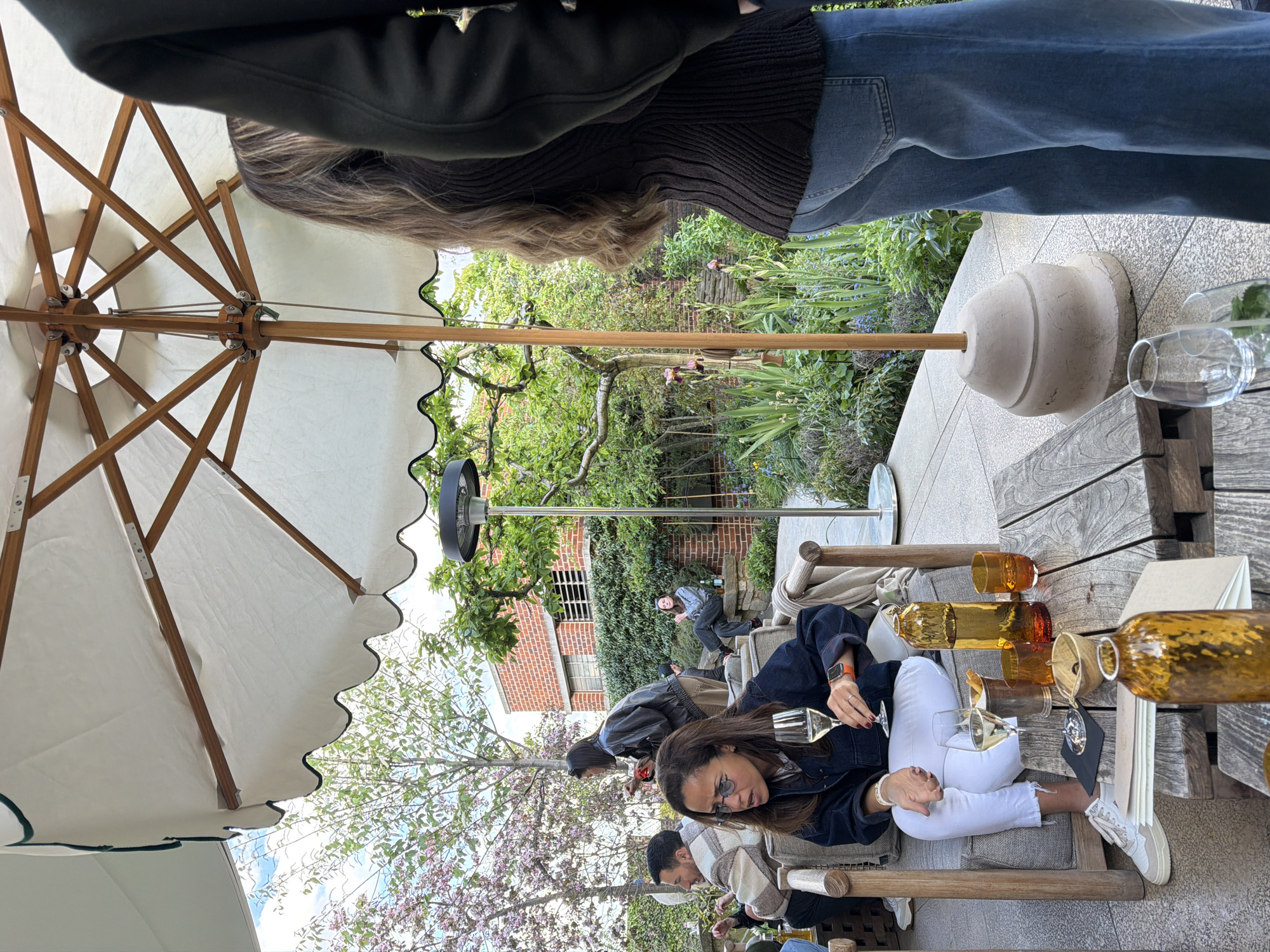 The image captures an outdoor dining scene in London, United Kingdom. Under a large white umbrella with a scalloped edge, patrons are seated at wooden tables. A woman wearing a denim jacket and white jeans is holding a wine glass and appears to be engaged in conversation. Other individuals are seated nearby, including one person in a beige and white plaid scarf.
On the table, there are multiple decorative amber-colored glass bottles, wine glasses, and a menu. In the background, a brick building covered in ivy is visible, along with flowering trees and other greenery. A person is seen walking through the foliage near the building. The foreground and background are blurred, suggesting a shallow depth of field.
