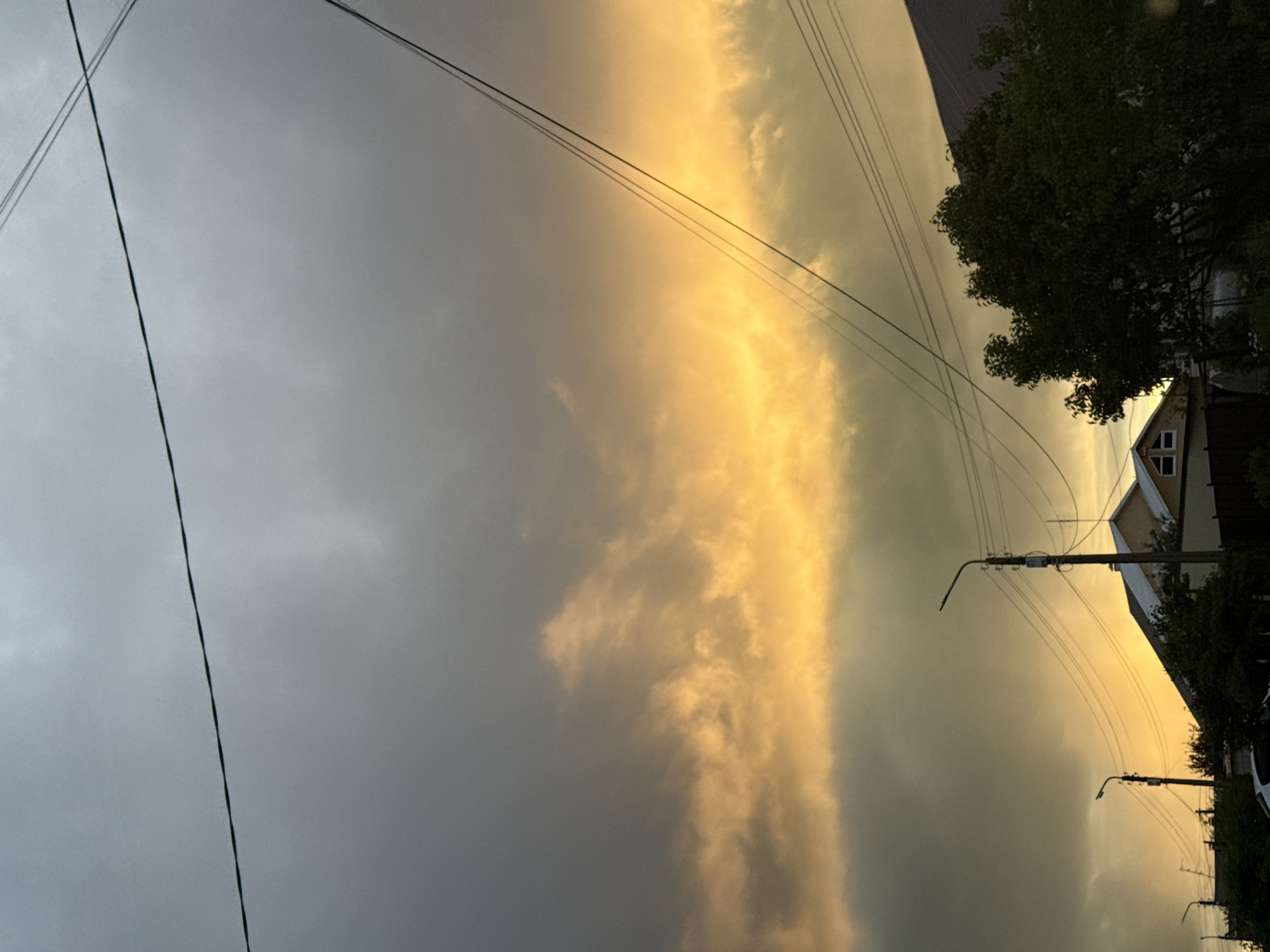 The image, rotated to depict the sky above a landscape, captures a dramatic moment of light and clouds. The sky is largely covered by heavy, dark gray clouds on the left and center, suggesting a recent or approaching weather event. This dark expanse is abruptly met by a vertical band of intensely bright, golden-orange clouds on the right, indicating direct illumination from a low sun, likely during sunset or sunrise.
Numerous dark utility lines crisscross the sky, extending from the upper left to the lower right. Several utility poles with streetlights are visible, particularly in the lower right and mid-right, supporting these lines and indicating a developed area. Below the dramatic sky and along the right side of the frame, elements of a residential landscape are present. Dark green foliage from trees and bushes obscures parts of the view, appearing heavily shadowed. A house with a pointed roof and visible windows can be seen on the far right, partially illuminated by the golden light. Further down, parts of another structure and a potential white vehicle are visible.
The scene depicts a residential area in Прикамский, Russia, characterized by its infrastructure and housing. The notable details include the stark contrast between the dark, overcast sky and the brilliantly lit golden clouds, suggesting a powerful weather phenomenon coinciding with either dawn or dusk. No visible text is present in the image.