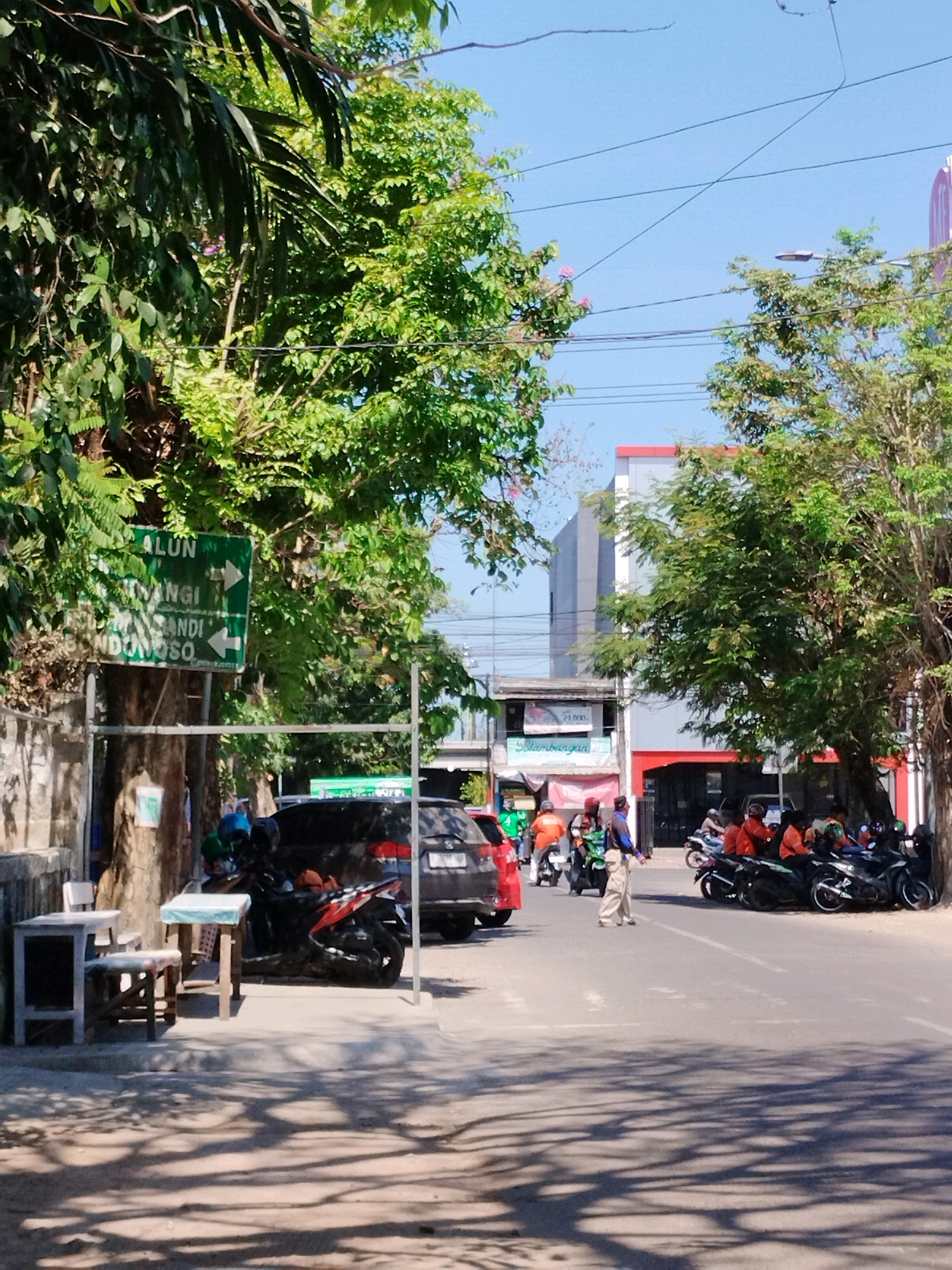 The image shows a street scene in Jember, Indonesia, on a sunny day. On the left side of the street, there is a green directional sign pointing to "ALUN", "WENGI", and "CANDI BONDOWOSO". Beside the sign, there are two small wooden tables with chairs. A black car is parked behind the tables. Several motorcycles are parked on the right side of the street, with some people wearing orange shirts standing near them. A person in a light-colored outfit is walking on the street. In the background, there are buildings and trees. The sky is clear and blue.