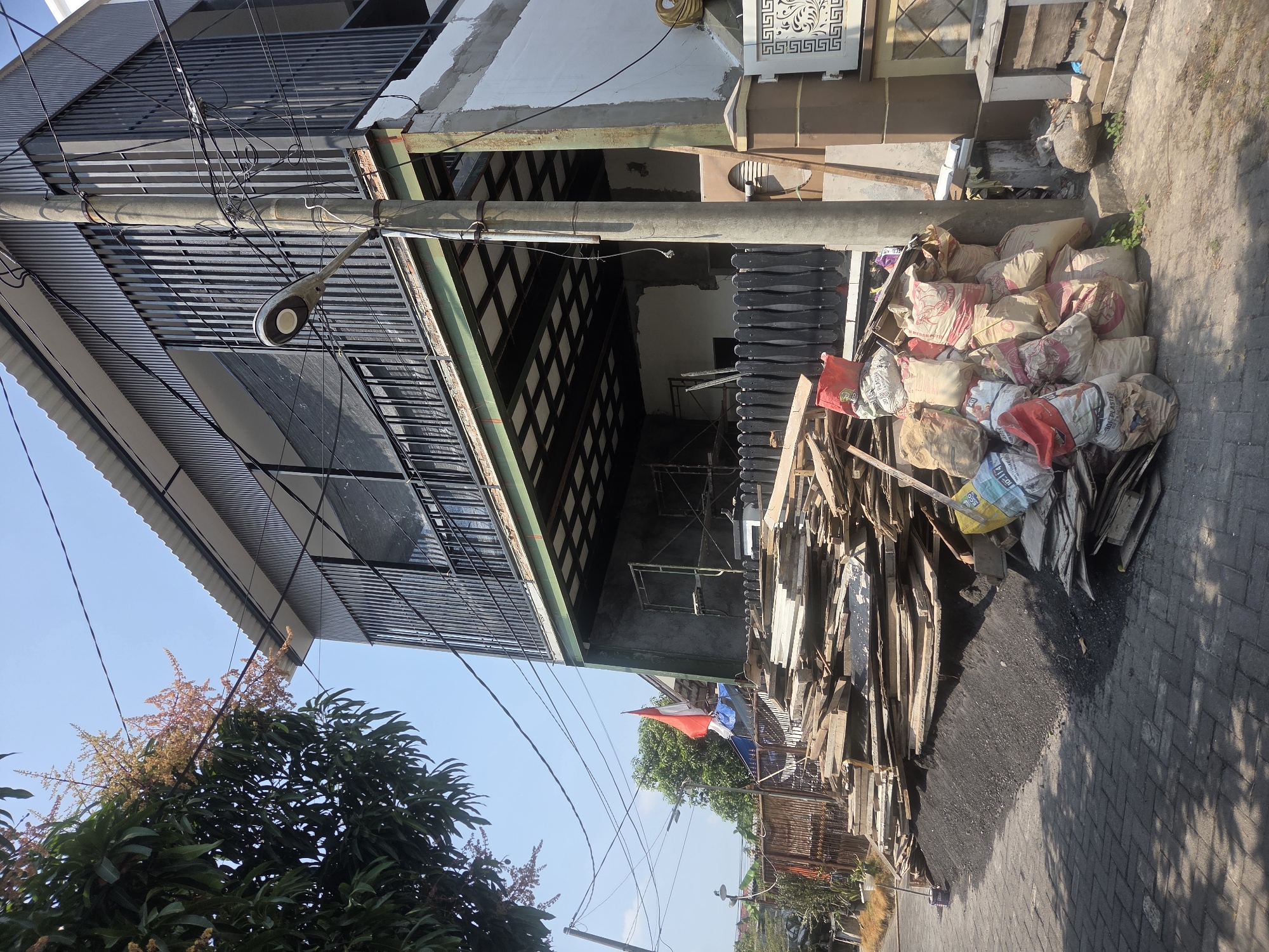 The image depicts a construction site in Sidoarjo, Indonesia, characterized by a partially constructed building and a large pile of building materials. The building, which appears to be two stories high, features a modern design with vertical metal siding on the upper level and concrete on the lower. A black metal railing encloses a balcony or walkway on the second floor. Scaffolding is visible on the lower level, suggesting ongoing construction work.
In the foreground, a substantial accumulation of construction debris is present. This includes a large stack of wooden planks and boards of various sizes, as well as numerous burlap sacks filled with what appears to be cement or plaster. Some of these sacks are red and white, indicating branding or contents. The debris is piled next to a gray, paved pathway that leads away from the building.
A tall concrete utility pole stands prominently in the midground, adorned with a street lamp and a tangle of electrical wires. To the left of the pole, an Indonesian flag is visible, fluttering against a clear blue sky. The surrounding environment suggests a moderately developed urban or suburban area, with trees and other structures visible in the background. The scene conveys a sense of ongoing development and the typical organized chaos of a construction project.