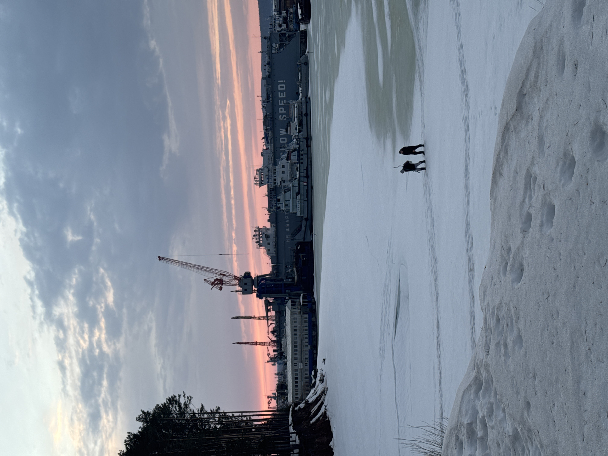 The image depicts a winter scene in the city of Chaikovsky, Russia. The view is oriented towards the Kama River, which is covered in snow and ice. Two individuals are walking on the ice surface. A shipyard is visible in the background, featuring several large ships with the phrase "LOW SPEED!" painted on one of them. Cranes are present at the shipyard. A forest line is seen on the left side of the image. The sky presents a gradient of colors from gray to pink, indicating either sunrise or sunset. Snow banks are visible on the right side of the image.