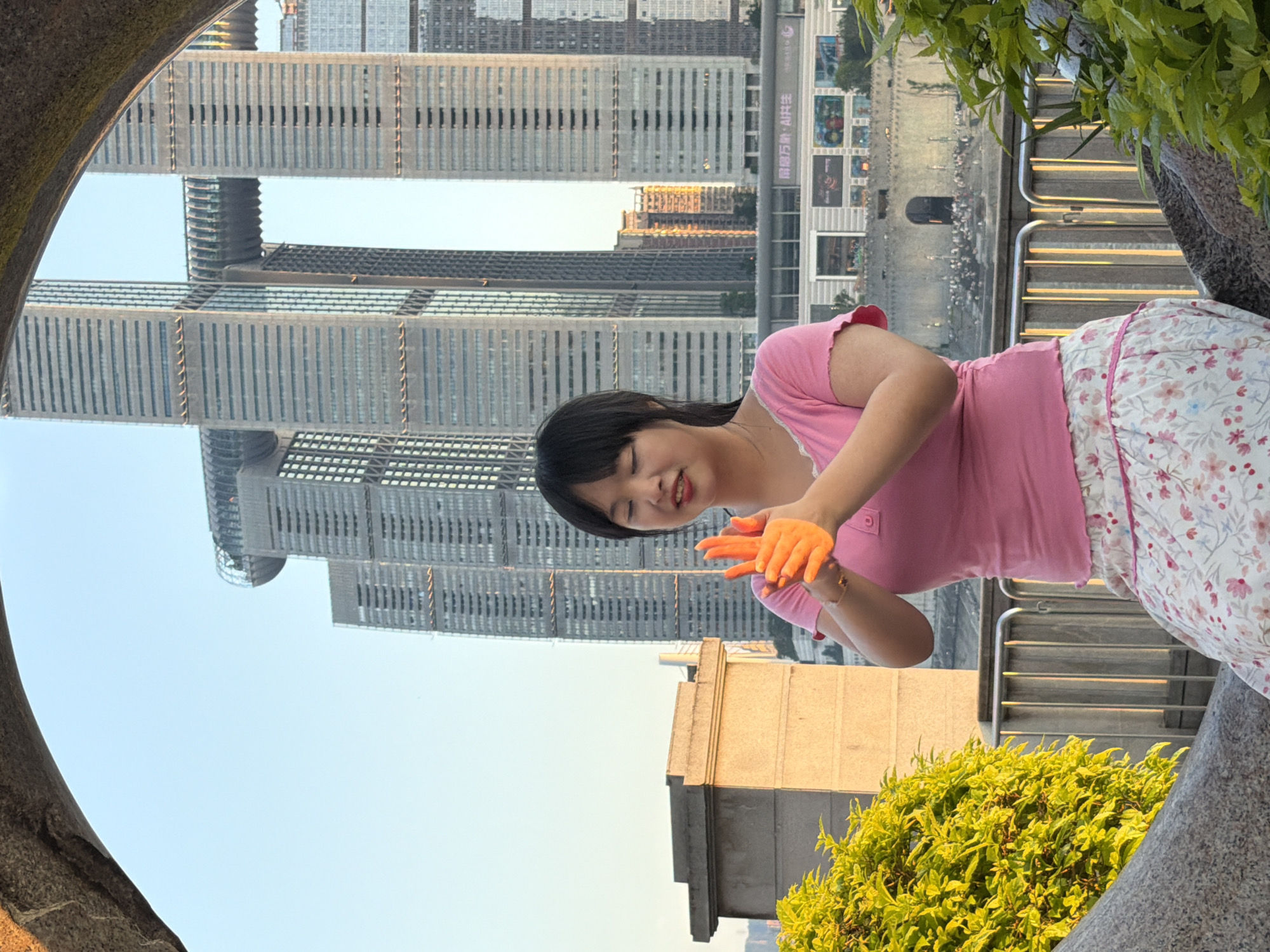 A young East Asian woman with dark hair and bangs is shown in a medium shot, seated outdoors and framed by a stone archway. She is wearing a pink t-shirt with lace trim and a floral skirt. Her hands are held in front of her chest, with her palms facing each other, and her fingers are illuminated by a warm, orange light. She is looking down at her hands with her eyes closed and a slight smile. In the background, the Raffles City Chongqing complex, a distinctive skyscraper complex featuring a horizontal skybridge connecting two towers, is visible against a clear blue sky. The buildings are tall and modern, with many windows. A concrete structure and lush green foliage are visible in the foreground to the left and right of the woman, respectively. A metal railing runs along a walkway behind her, and a wide river with numerous birds on its banks can be seen further in the distance. The lighting suggests it is late afternoon or early evening, with a warm glow cast on the scene. The overall impression is one of peaceful contemplation in an urban setting.