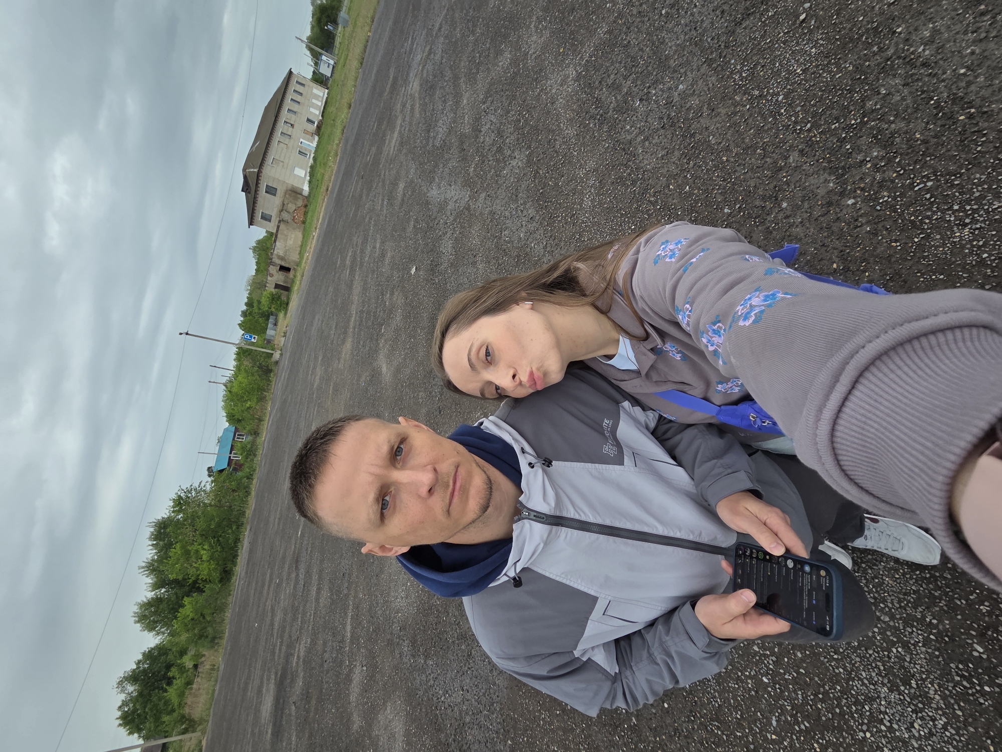 This media file captures an outdoor selfie of a man and a woman, likely taken in Nizhneleninskoye, Russia. The photograph has a diagonal orientation, with the camera appearing to be held by the woman.
In the foreground is a man, likely in his 30s-40s, with short hair, a light beard, and blue eyes. He is wearing a two-tone grey and light grey hooded jacket, on which the word "ROUTE" is partially visible. He looks directly at the camera with a neutral expression, holding a black smartphone that displays a dark-themed messaging application with various chat entries, though the specific text is unreadable.
Resting her head affectionately on the man's shoulder is the woman, who has long brown hair. She faces the camera with a playful, puckered-lips expression. She is dressed in a grey hooded top adorned with light blue floral patterns. A blue object, possibly a bag, is partially visible near her arm.
They are positioned on a dark, textured ground composed of small pebbles, resembling a gravel or unpaved area. The sky is entirely overcast, indicating a cloudy day with no visible direct sunlight. In the background, there are various environmental elements: a large, multi-story building constructed of light grey blocks, possibly an older apartment or commercial structure, stands to the left side of the frame. Some of its windows appear broken or boarded up. Further into the distance, a small house with a distinct blue roof is visible nestled among lush green trees. A utility pole and a blue road sign, which appears to be a parking "P" symbol, are also discernible. The overall scene suggests a somewhat aged or developing outdoor environment.