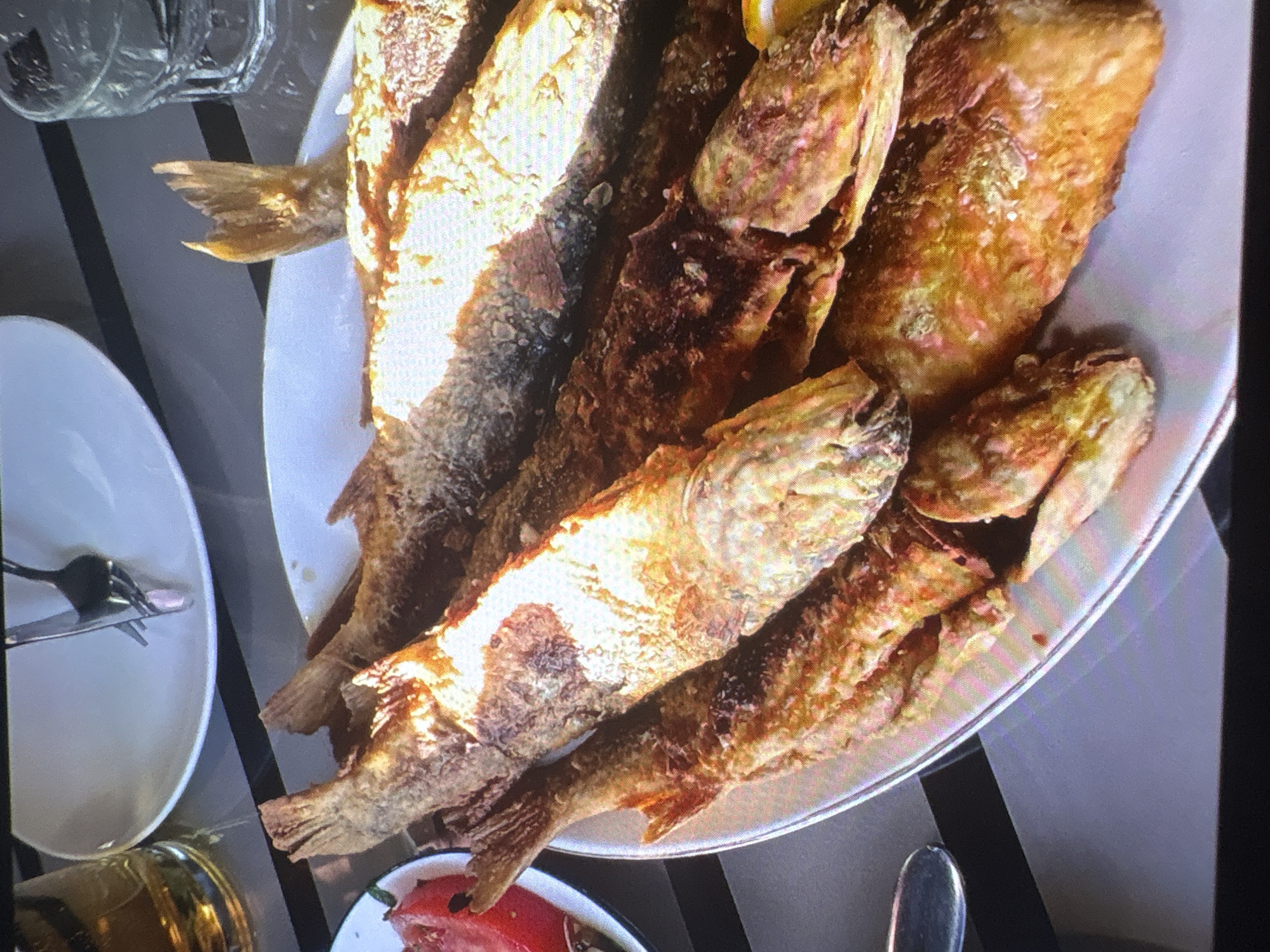In Batumi, Georgia, a white plate piled high with fried fish is the focal point of the image. The fish, various sizes and golden brown, are arranged in a layered fashion, with some overlapping others. Their tails and fins are visible, suggesting they are whole fish. The plate is set on a dark surface, likely a table, which is reflected in the polished surface. To the left of the plate, a white dish with what appears to be slices of red fruit, possibly tomatoes or watermelon, is partially visible. Behind the plate, a white plate with cutlery, a glass of amber liquid, and another glass with a clear liquid and ice can be seen, indicating a meal setting. The lighting is bright, casting shadows and highlighting the textures of the food. The overall scene suggests a casual dining experience in Batumi.