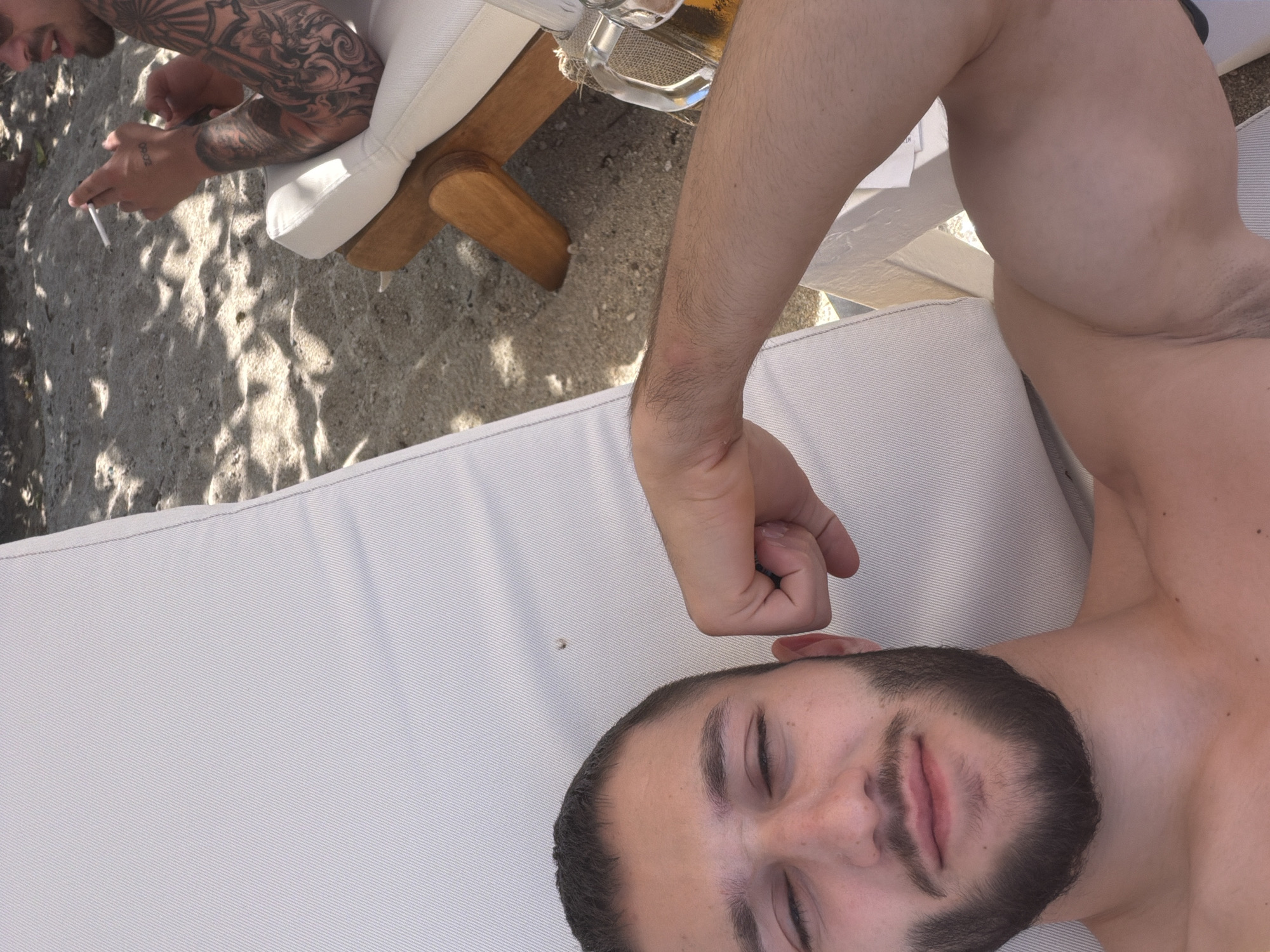 A medium, high-angle shot depicts a man with a beard lying on a white lounge chair at a beachside location in the Pallini Municipal Unit, Greece. The man has his eyes closed and appears to be resting. Another man, partially visible, with a tattoo on his arm is seated nearby, holding a cigarette. A glass mug of beer is placed on a small wooden table beside the lounge chair. Sandy ground and foliage are visible in the background. The scene is illuminated by natural daylight.