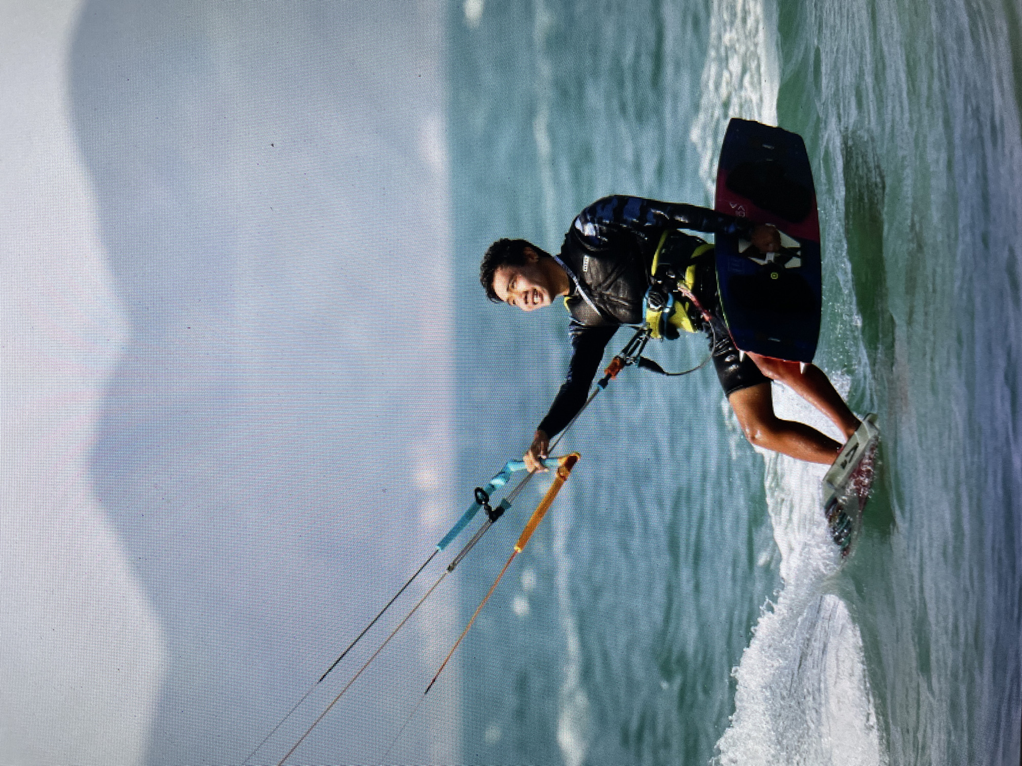 A man is kitesurfing in Bacong, Philippines. He is wearing a black wetsuit and a harness, holding onto a kite bar with his hands. He is riding a kiteboard on the water. The water is choppy, with whitecaps visible. The sky is overcast.