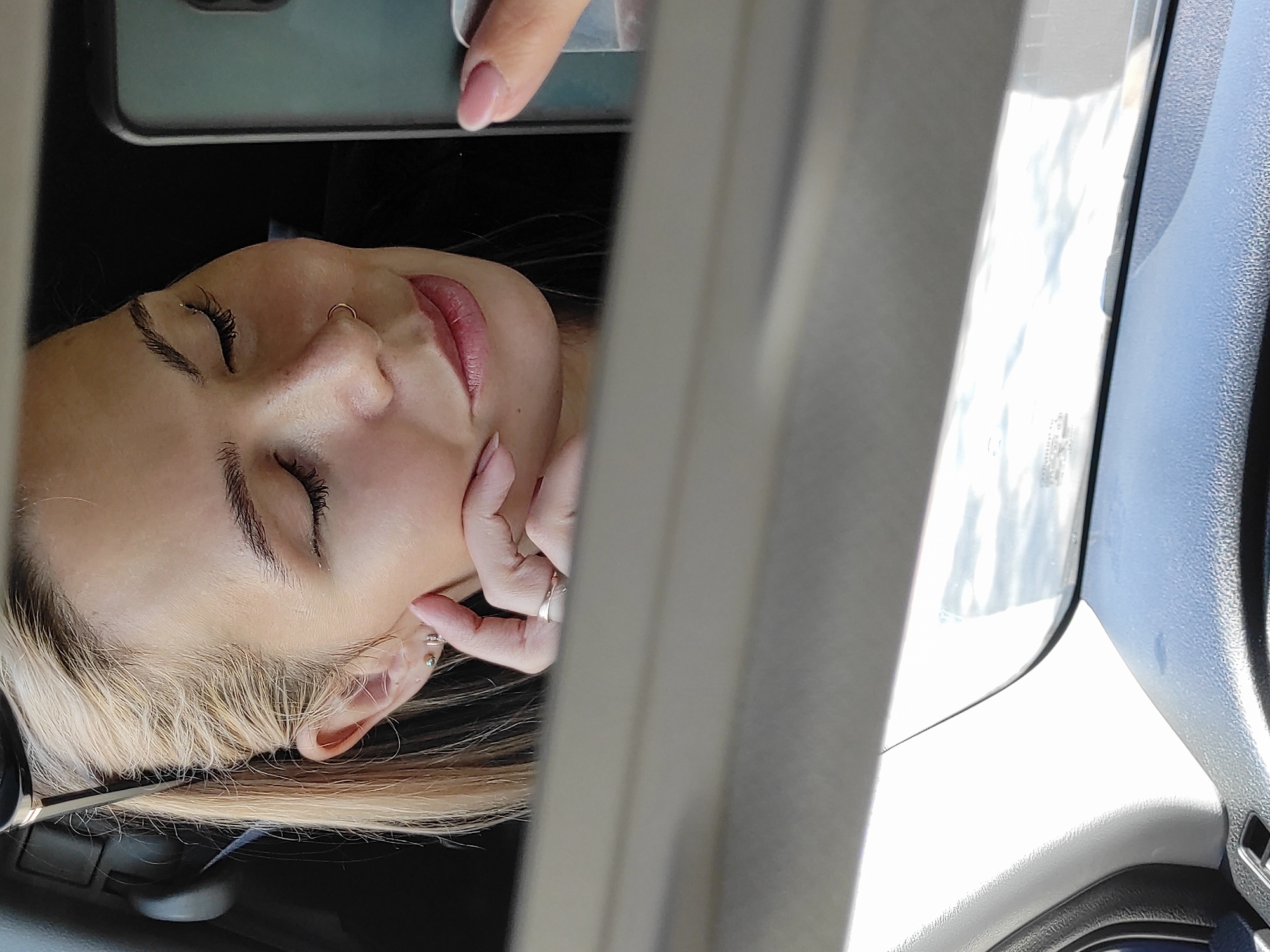 A woman with light blonde hair and fair skin is depicted inside a car. She has her eyes closed and is touching her face with her fingers. She has a nose ring and multiple earrings. The interior of the car is visible, including part of the dashboard and window. The scene is in the city of Chaikovsky, Russia.