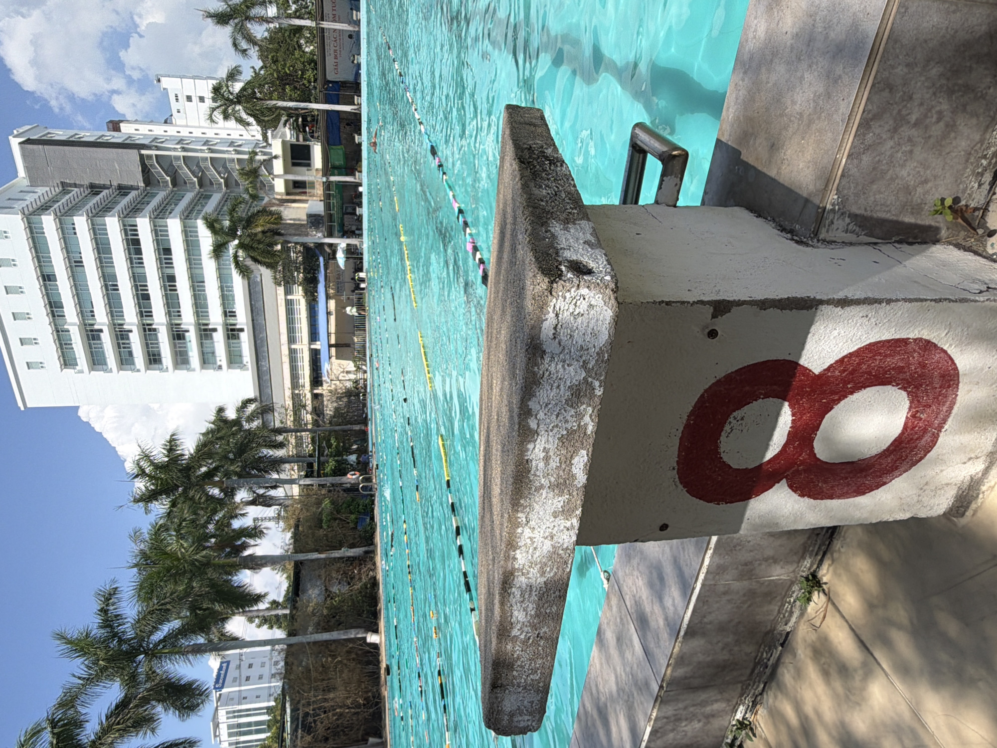 An outdoor swimming pool scene is depicted on a bright, sunny day, characteristic of a coastal or resort city like Nha Trang, Vietnam. The viewpoint is low, looking across the pool from near a concrete starting block. This block prominently displays a large red number "8" and shows signs of weathering and use. Beside it, a silver metal handrail extends into the clear, inviting turquoise water.
The swimming pool is filled with clear, light blue water, and multiple lanes are defined by floating lines of buoys, suggesting it's designed for lap swimming or competitions. At least one person can be seen swimming in one of the lanes.
In the mid-ground and background, several tall, modern white buildings with numerous windows rise against a clear blue sky dotted with fluffy white clouds. These structures likely represent hotels or apartment complexes. Numerous lush green palm trees are scattered around the pool area and among the buildings, enhancing the tropical atmosphere.
A banner in the background features Vietnamese text, partially visible, which reads "GIẢI BƠI CÁC NHÓM TUỔI," translating to "Swimming Competition for Age Groups." Small green plants are observed growing from cracks in the concrete around the pool's edge. The strong sunlight casts distinct shadows, confirming it is daytime.