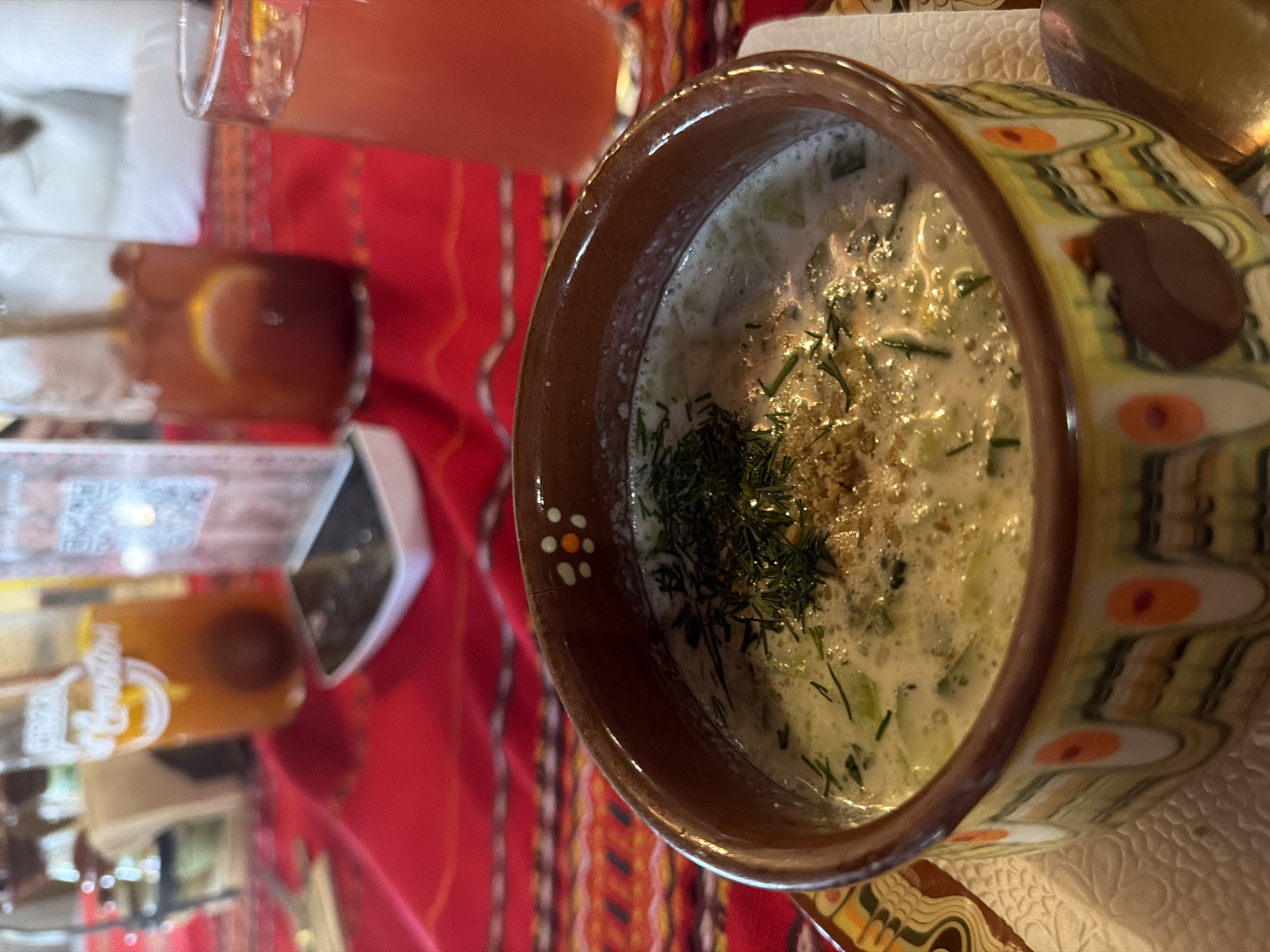 The image presents a bowl of Tarator, a cold cucumber soup, placed on a colorful woven tablecloth. The bowl features a brown rim and a decorative pattern of olive, orange, and brown shapes. Dill and what appears to be ground nuts garnish the soup. In the background are a glass of juice and another beverage with a lemon slice. The photograph appears to be taken from above. The setting is in Panagyurishte, Bulgaria.