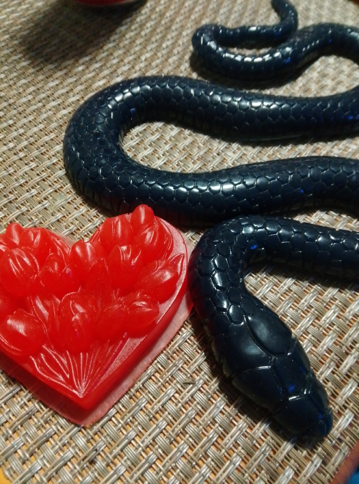 A black, coiled snake figure with a segmented texture rests on a woven mat next to a red heart-shaped object with carved floral details. The snake is in the foreground and takes up the majority of the frame. The heart is to the left of the snake. The image is a close-up shot, and the background is out of focus. There are no people or any other recognizable objects in the scene. The location in the city of Wedel, Germany, is not visible or discernible from the image.