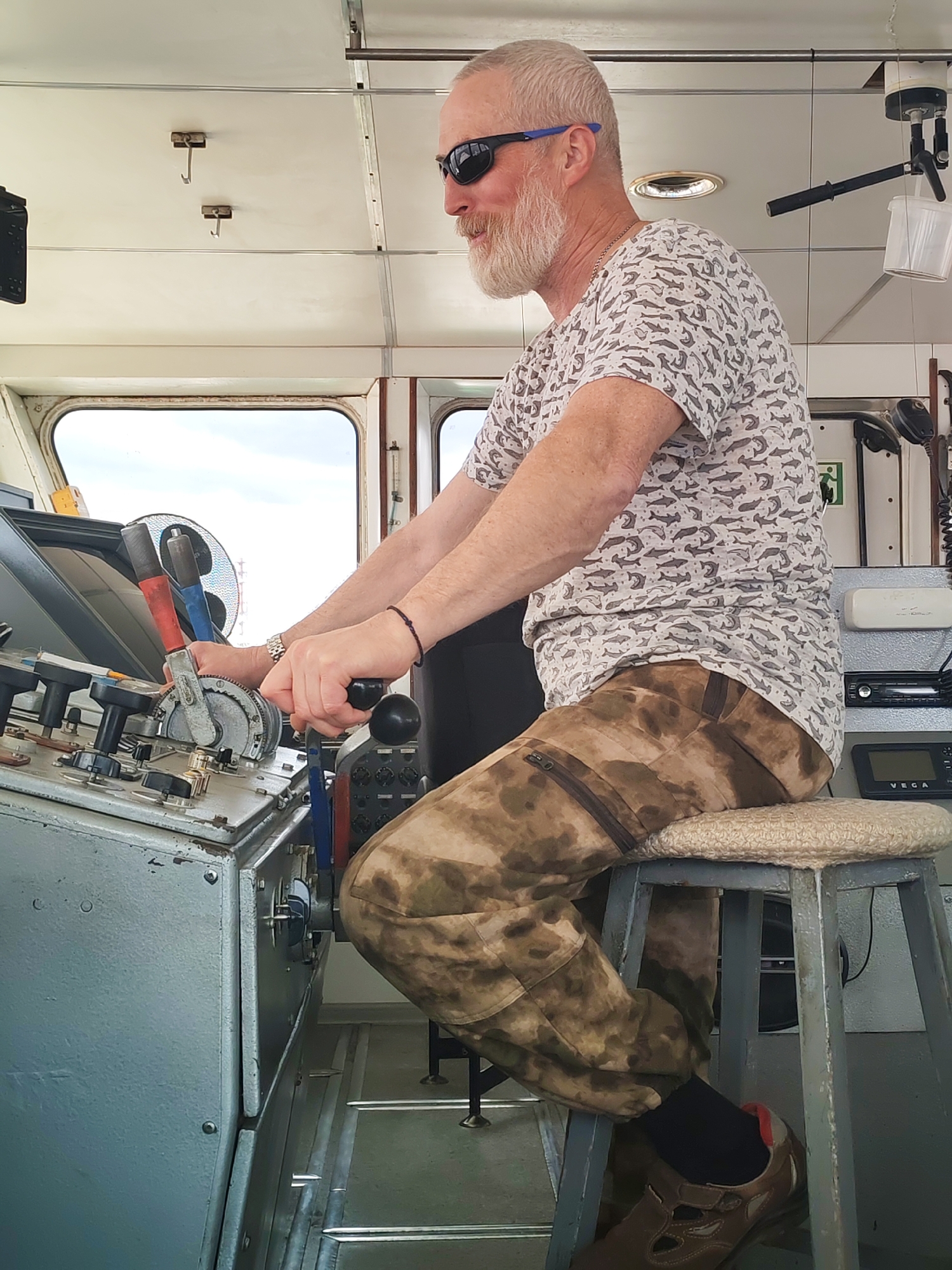 The image shows a man operating controls inside what appears to be the bridge or control cabin of a marine vessel, likely during the daytime. He is a middle-aged to elderly man with light-colored (possibly gray or white) short hair, a prominent beard, and a mustache. He wears dark sunglasses with blue accents, a light-colored t-shirt patterned with dark fish or sharks, and camouflage-style cargo pants.
The man is seated on a simple grey metal stool with a beige, woven cushion. His hands are actively engaged with the control panel in front of him. His right hand grips a black lever, while his left hand operates a set of levers, one red and one blue, which are part of a larger, grey metallic console filled with various dials, buttons, and switches.
Through the large windows in the background, a bright sky and a hazy, distant view of what might be land or industrial structures are visible. To the right of the man, a small electronic device is visible with the brand name "VEGA" printed on it, positioned below a car stereo unit. An arm extends from the ceiling above this equipment, holding a clear plastic container. Two metal hooks are also visible on the ceiling. Partially visible on the wall behind the man's left shoulder is a green emergency exit symbol. The man appears focused on the task of operating the vessel. He is wearing a chain necklace, a dark bracelet on his right wrist, dark socks, and brown shoes.