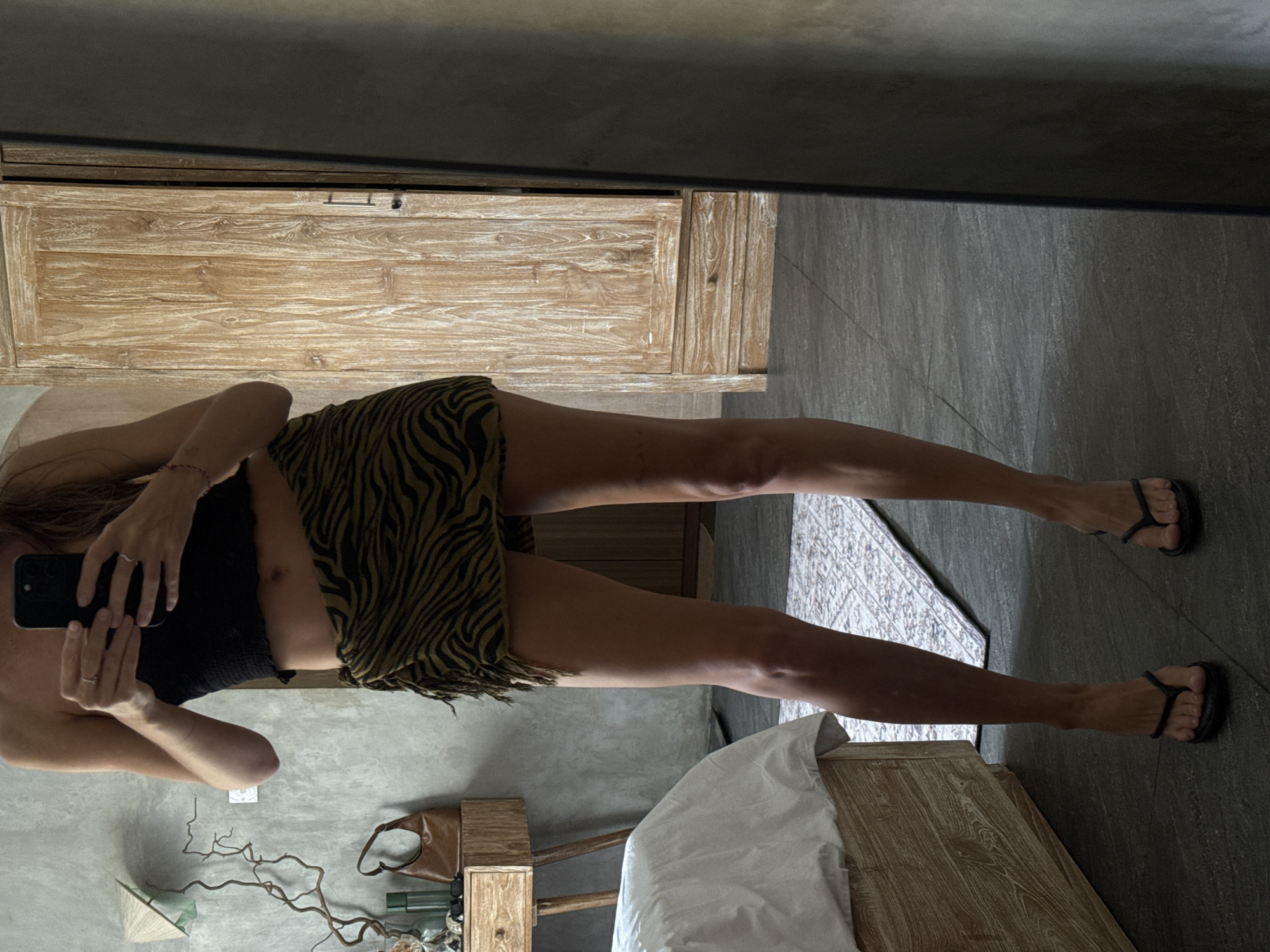 A woman in a black top and zebra-striped skirt is taking a mirror selfie in a room in North Kuta, Indonesia. The room contains a wooden dresser, a bed with white sheets, a small wooden table with a brown purse, a vase with branches, a conical hat, and other small objects. The floor is tiled. The woman is wearing black flip-flops. A rug is visible on the floor in the mirror's reflection.