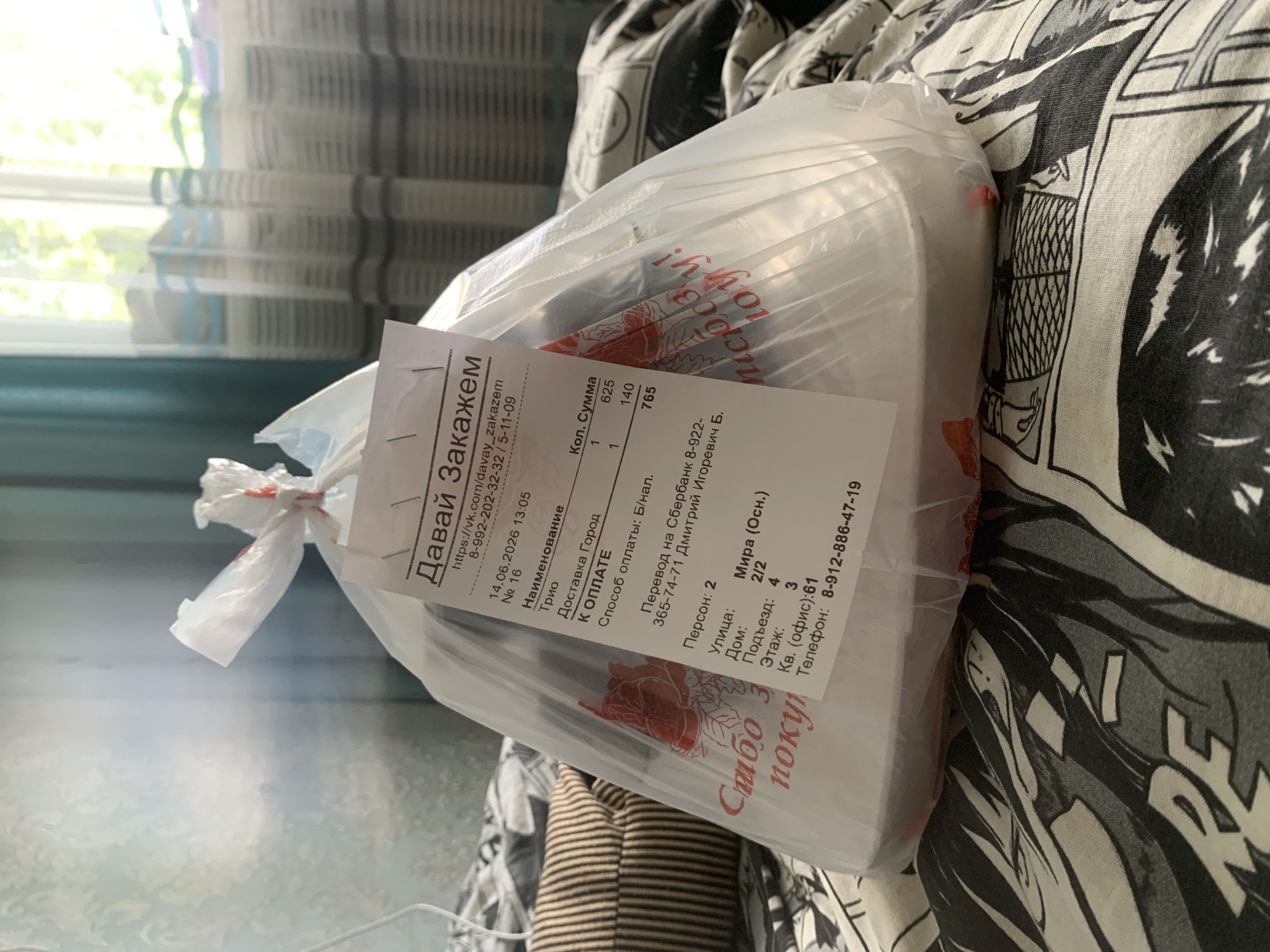 A white plastic bag containing food is resting on a bed with a black and white patterned comforter. A white receipt is attached to the bag by staples. The receipt displays text in Russian, including "Давай Закажем" (Let's Order), a website address "https://vk.com/davay_zakazem", and a phone number "8-992-202-32-32". It also shows a date "14.06.2026 13:05", an order number "№ 16", item names like "Трио" (Trio) and "Доставка Город" (City Delivery), quantities and prices, and delivery address details. The address includes "Мира (осн.)" (Mira (main)), "Улица: Мира", "Дом: 2/2", "Подъезд: 4", "Этаж: 3", "Кв.(офис):61", and a phone number "8-912-886-47-19". The bag has a red floral design printed on it, and the phrase "Спасибо за покупку!" (Thank you for your purchase!) is visible. The background shows a window with light blue and white striped curtains, suggesting it is daytime. The bed is positioned near a wall with a light blue textured wallpaper.