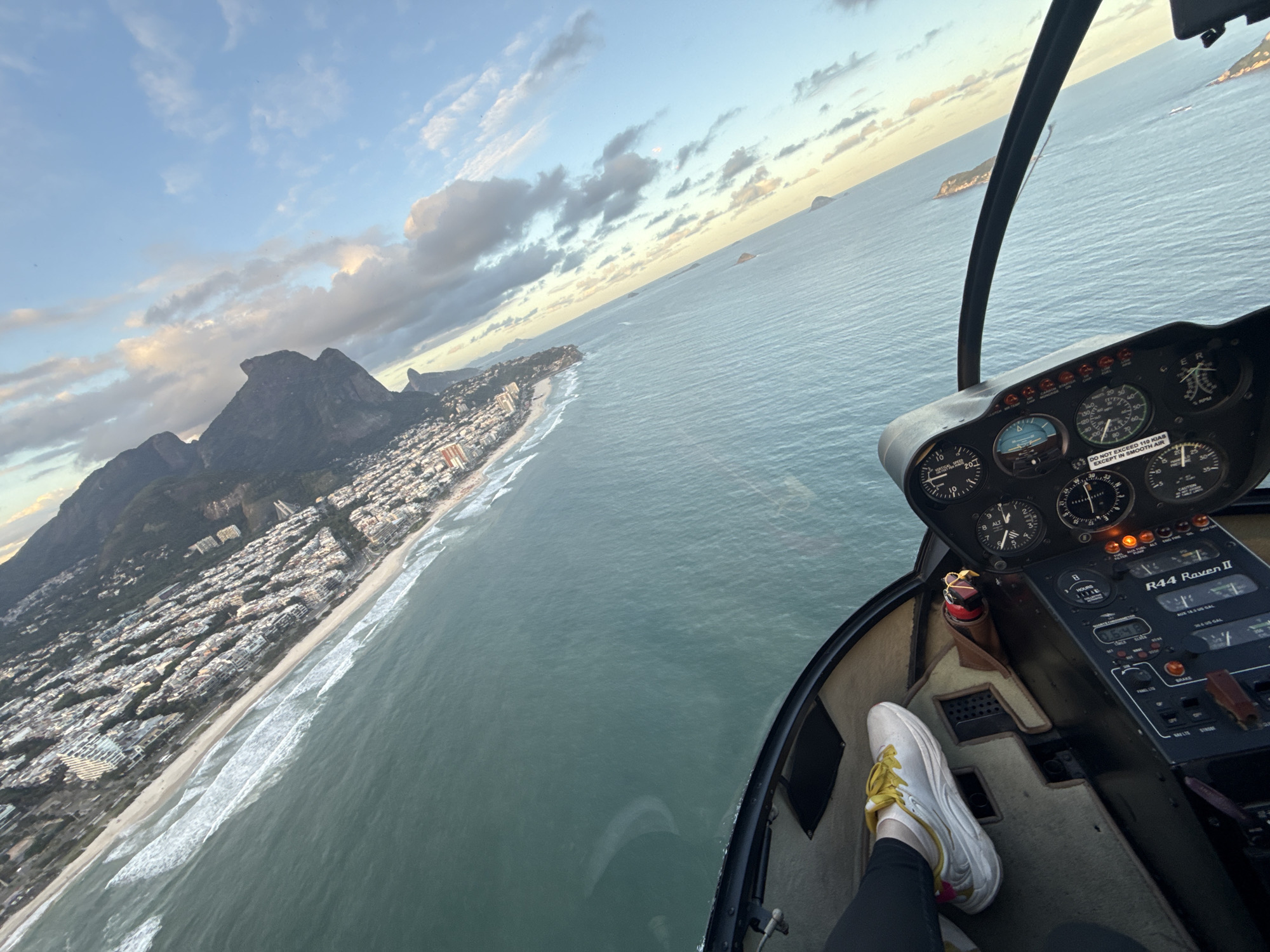 This is an aerial photograph captured from a Robinson R44 Raven II helicopter, looking out over the coastline of Rio de Janeiro, Brazil. The scene depicts the western part of the city, specifically overlooking the São Conrado beach and urban area, with the prominent Pedra da Gávea mountain visible on the left side of the frame.
The image is taken from a high vantage point, with the helicopter's interior forming the right and bottom foreground. A person's left foot, clad in a white sneaker with yellow laces and accents, is visible on the carpeted floor of the cabin, positioned near the control panel. The instrument panel itself is black with various analog dials, digital displays, and illuminated indicator lights. A warning label on the panel states, "DO NOT EXCEED 110 KIAS EXCEPT IN SMOOTH AIR." A red-capped emergency switch is visible in a holder below the panel.
The main subject of the landscape is the long, light-colored sandy beach, stretching diagonally across the mid-ground. White waves break consistently along the shore, meeting the turquoise-green ocean. Immediately behind the beach, a dense urban residential area with numerous white and light-colored buildings extends towards the base of a series of large, dark, tree-covered mountains. The most distinctive mountain features a flat, cliff-like summit.
The vast expanse of the Atlantic Ocean occupies the right half of the image, appearing calm with a slight reflection of the sky. Several small, rocky, dark islands are scattered offshore, receding towards the horizon. The sky is partly cloudy, with a mix of clear blue sky and scattered white and grey cumulus clouds. The lighting suggests either early morning or late afternoon, with a soft, warm glow visible near the horizon.