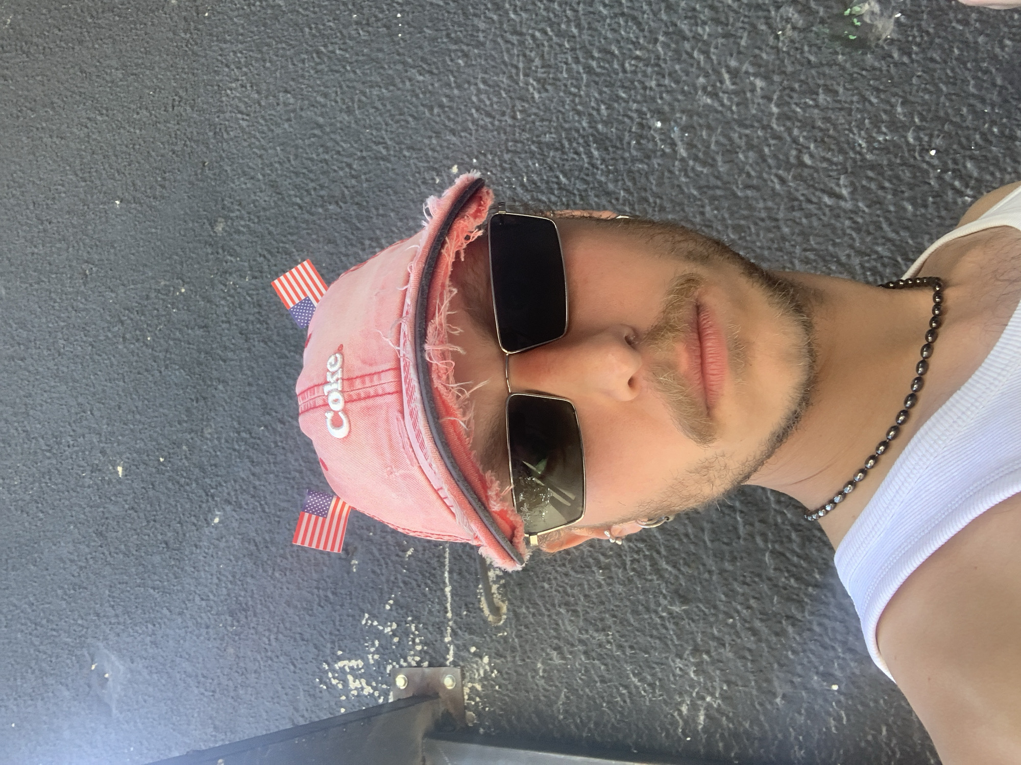 An individual is depicted in a head-and-shoulders portrait, facing the camera directly, positioned outdoors in Tel-Aviv, Israel. The subject, appearing male, has light skin, a light brown mustache, beard, and sideburns. They wear a distressed, faded red baseball cap featuring "Coke" embroidered in white on the front, flanked by two small American flag pins. Rectangular Ray-Ban sunglasses with dark lenses and thin silver frames cover the eyes. The individual has multiple silver earrings, including hoops on both ears and an additional stud/hoop on the left ear. A white tank top and a dark, beaded choker-style necklace are worn. The background is a dark, textured wall, likely painted concrete, exhibiting a coarse, mottled surface. A dark vertical metal beam or fixture is secured to the wall on the left, evidenced by two visible silver bolts. The scene is brightly illuminated by direct daylight.