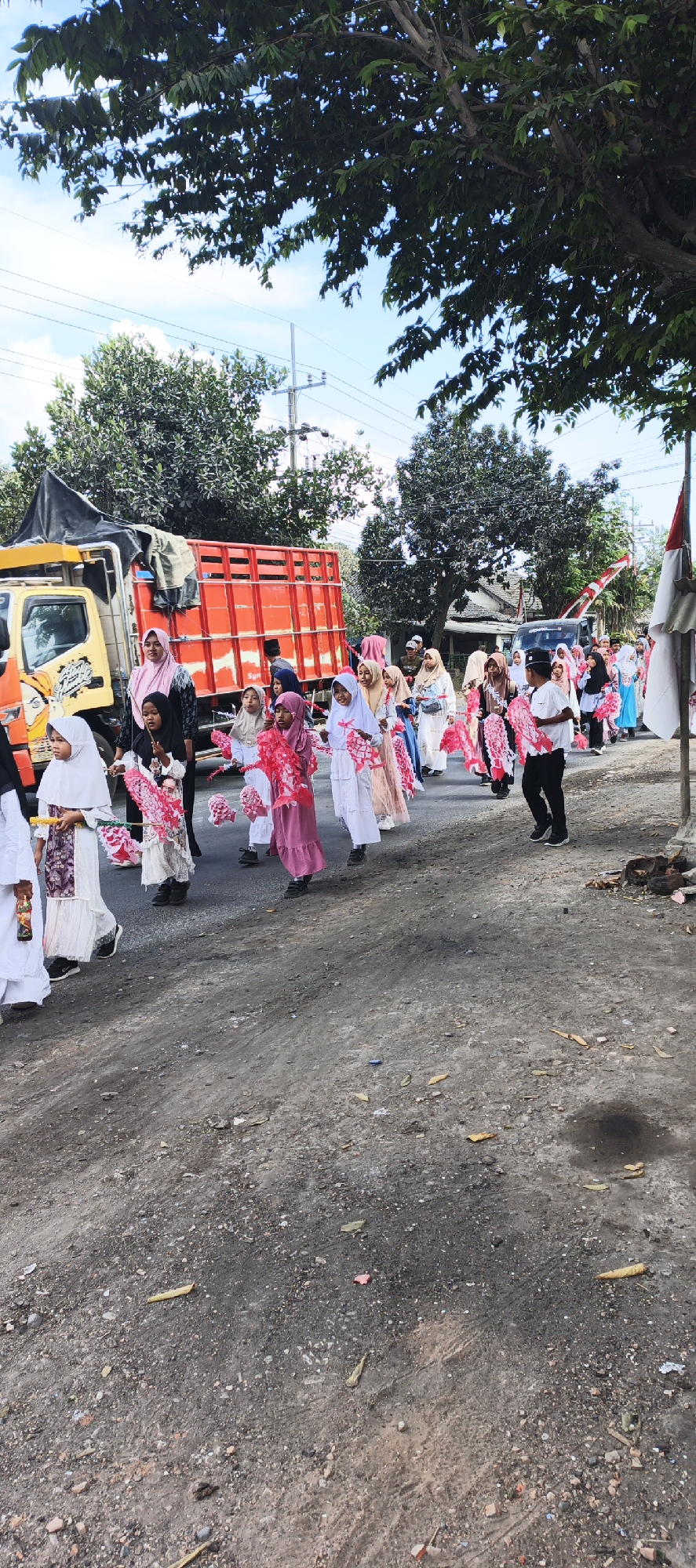 In Grenden, Indonesia, a group of children, predominantly girls dressed in hijabs and modest clothing, are participating in a street procession. They are holding colorful, possibly paper or fabric, decorations, predominantly in shades of pink. The procession is moving along a dusty, unpaved road, with a large orange and red truck on the left side of the image. Trees line the right side of the road, and power lines are visible overhead, suggesting an outdoor, possibly rural or semi-urban setting. Several adult figures, also in traditional attire, are interspersed within the group of children, seemingly supervising or participating in the event. The overall scene depicts a cultural or religious celebration.