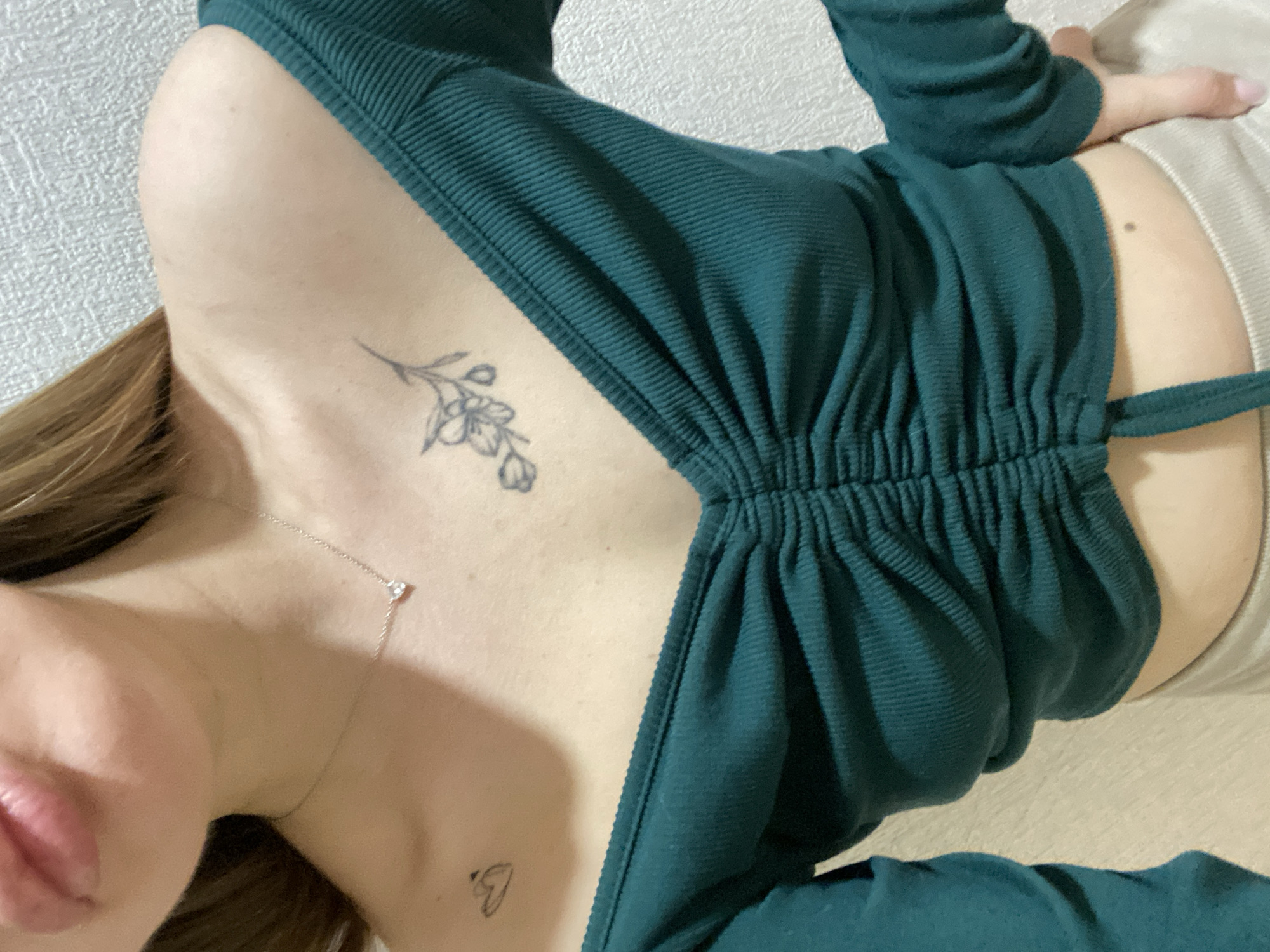 The image is a close-up, angled shot of a person wearing a green, ribbed top. The person has fair skin and is wearing a delicate necklace. Tattoos are visible on the chest area: a small floral design near the collarbone and a tiny heart symbol lower down. The person's lips are visible in the lower left corner of the frame. The background is a light, neutral color. The location is not discernible from the image and there is no indication that the image was taken in Izhevsk, Russia.