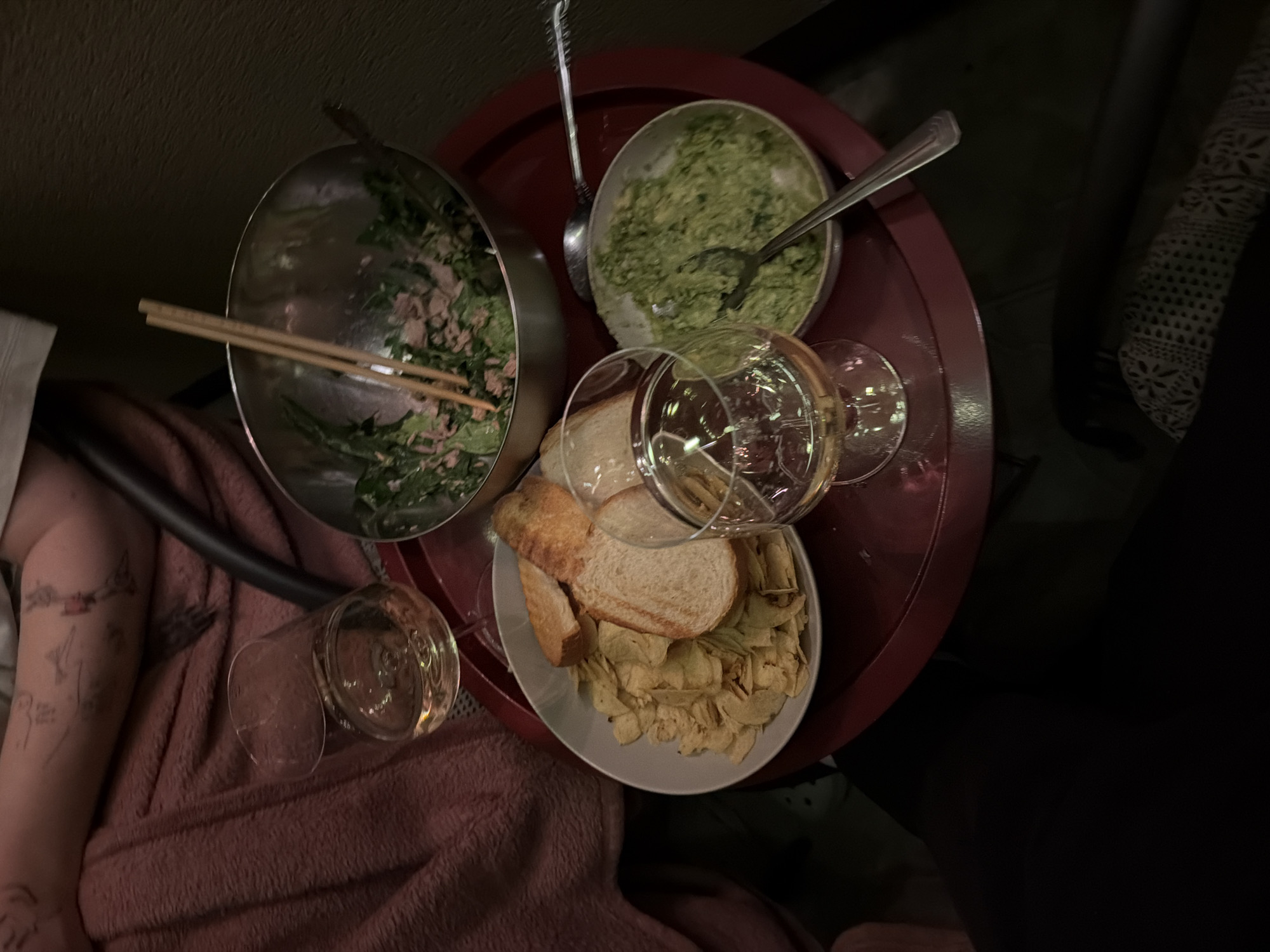 A top-down, dimly lit, indoor shot captures a red tray on a dark surface, holding a meal and drinks. The tray contains a stainless steel bowl with salad and chopsticks, a white bowl with potato chips and bread slices, and two glasses of white wine. A small bowl filled with a green dip, possibly guacamole, sits on the tray with a spoon. A person's arm, adorned with tattoos, rests on a pink blanket to the left of the tray. The overall scene suggests a casual dining setting, possibly in an apartment within the Zvezdara Urban Municipality of Serbia.