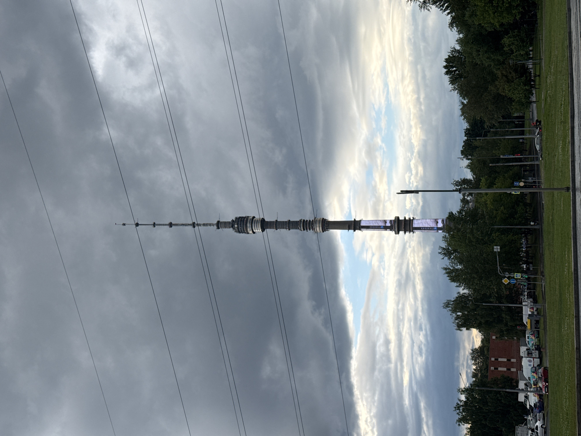 This is a photograph of a cityscape with a prominent television tower. The tower is tall and slender, with a distinctive design featuring multiple levels and a bright, illuminated section near the top. Several parallel power lines crisscross the frame in the foreground, obscuring parts of the sky and the tower.
The sky is dramatic, with a mix of dark, heavy storm clouds on the left and lighter, brighter clouds with patches of blue sky on the right, suggesting a weather front moving through. This creates a contrast between a darker, more ominous atmosphere on one side and a brighter, more open feel on the other.
Below the tower and to the right, a line of trees borders a grassy area. Beyond the trees, there is a street with several parked vehicles, including trucks and cars. Traffic lights and road signs are visible, indicating an urban environment. A red brick building can be seen in the background.
The overall scene appears to be a daytime shot, likely late afternoon or early evening given the lighting conditions and cloud formations. There are no visible people in the frame. The weather appears to be changing, with the transition from dark clouds to brighter ones.
The image is oriented vertically. The location is identified as Moscow, Russia. There is some text visible on the illuminated section of the tower, but it is too blurry to read clearly. The contrast in the sky and the presence of the imposing tower give the image a somewhat dramatic and imposing feel.