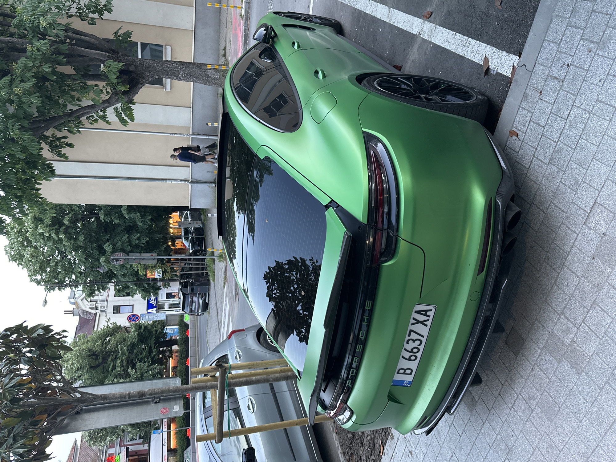 A green Porsche Panamera is parked in Varna, Bulgaria, alongside a street with buildings and trees. The car's license plate is B 6637 XA. A silver-colored car is parked directly in front of the Porsche. Two people are walking in the background near a light-colored building. The street features signs and pedestrian crosswalks.