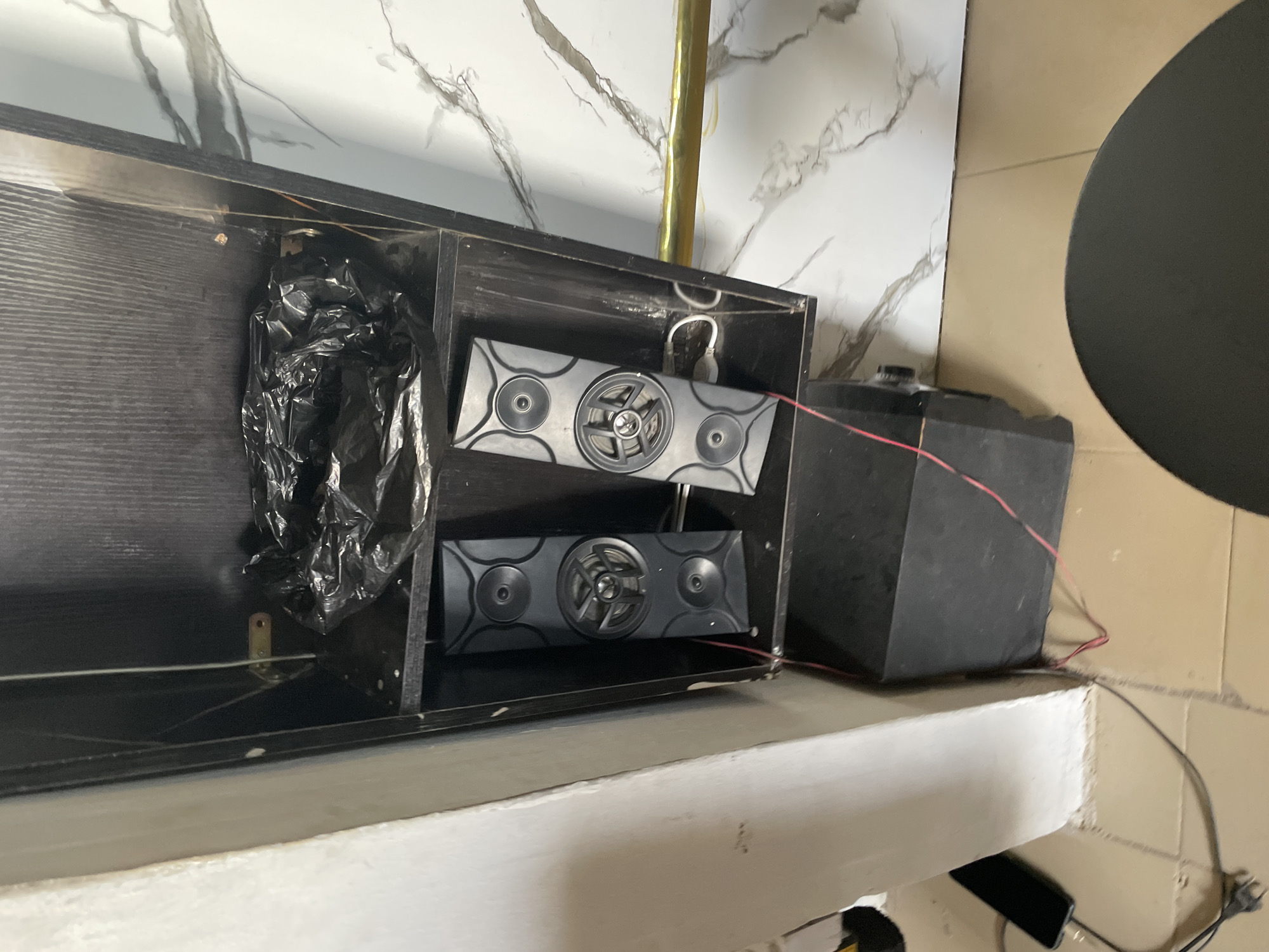 In Jalingo, Nigeria, within a dimly lit room, a dark, narrow cabinet stands against a wall with a marble-patterned finish. The cabinet's interior is divided into shelves. On the top shelf, a crumpled black plastic bag rests. Below it, two tall, black speakers are positioned vertically. Each speaker has a central circular speaker driver surrounded by four smaller circular elements. A black subwoofer is located on the floor directly beneath the speakers, connected to them by thin red wires. A power cord for a phone charger is visible on the floor to the left of the cabinet, plugged into an outlet and leading to a black smartphone. A golden rod or railing is mounted horizontally on the right side of the cabinet. The floor is tiled with a beige and brown checkered pattern.